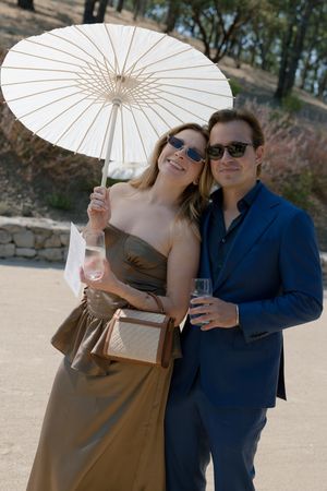 A couple in formal attire poses with a white parasol and drinks at an outdoor event.