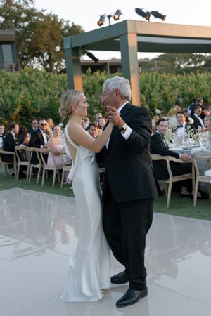 A couple performs their first dance at an outdoor wedding reception on a white dance floor under a modern pergola structure.
