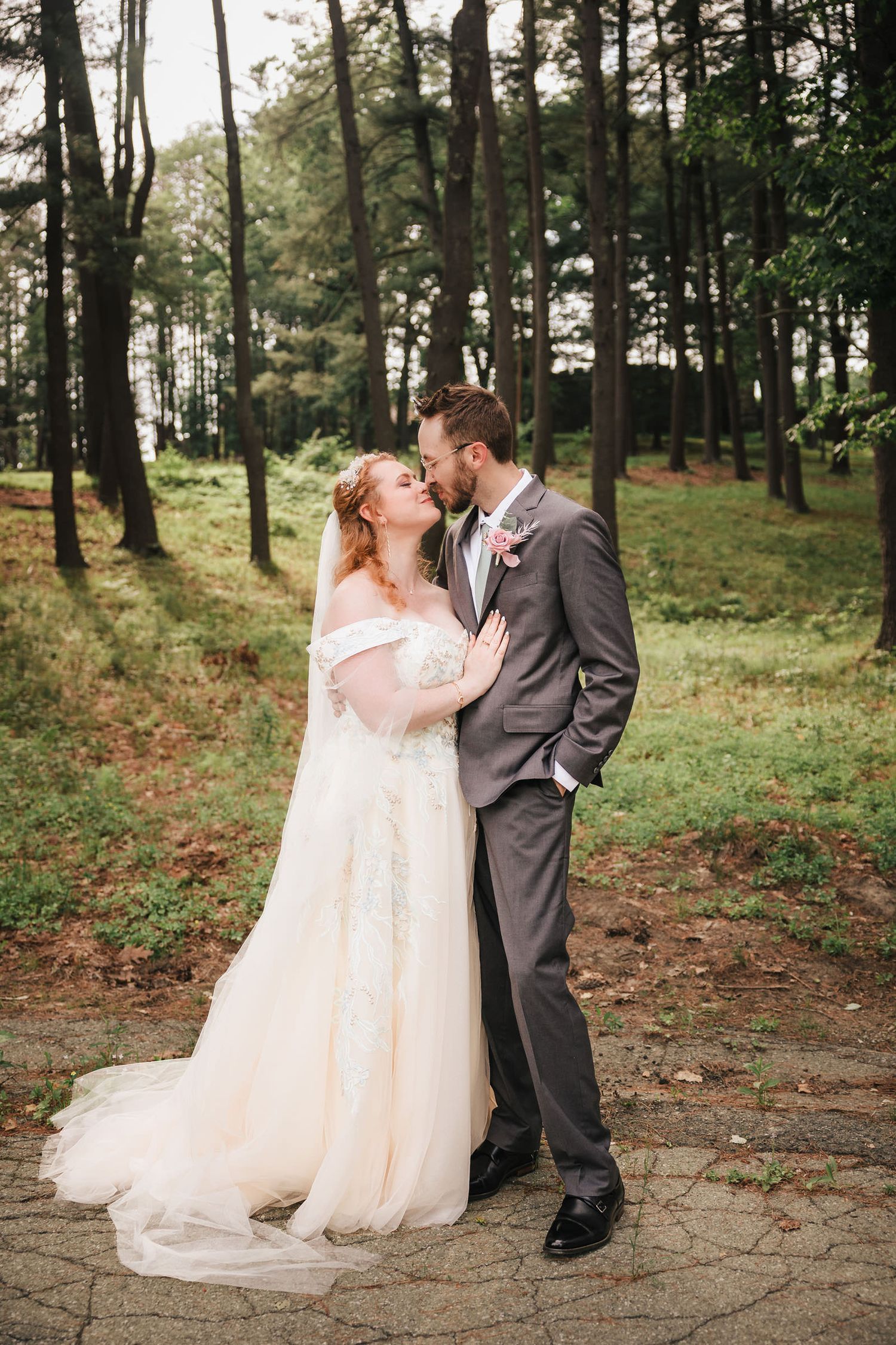 Bride and groom touching foreheads in a sunlit pine forest, her wedding dress train pooling on the ground.