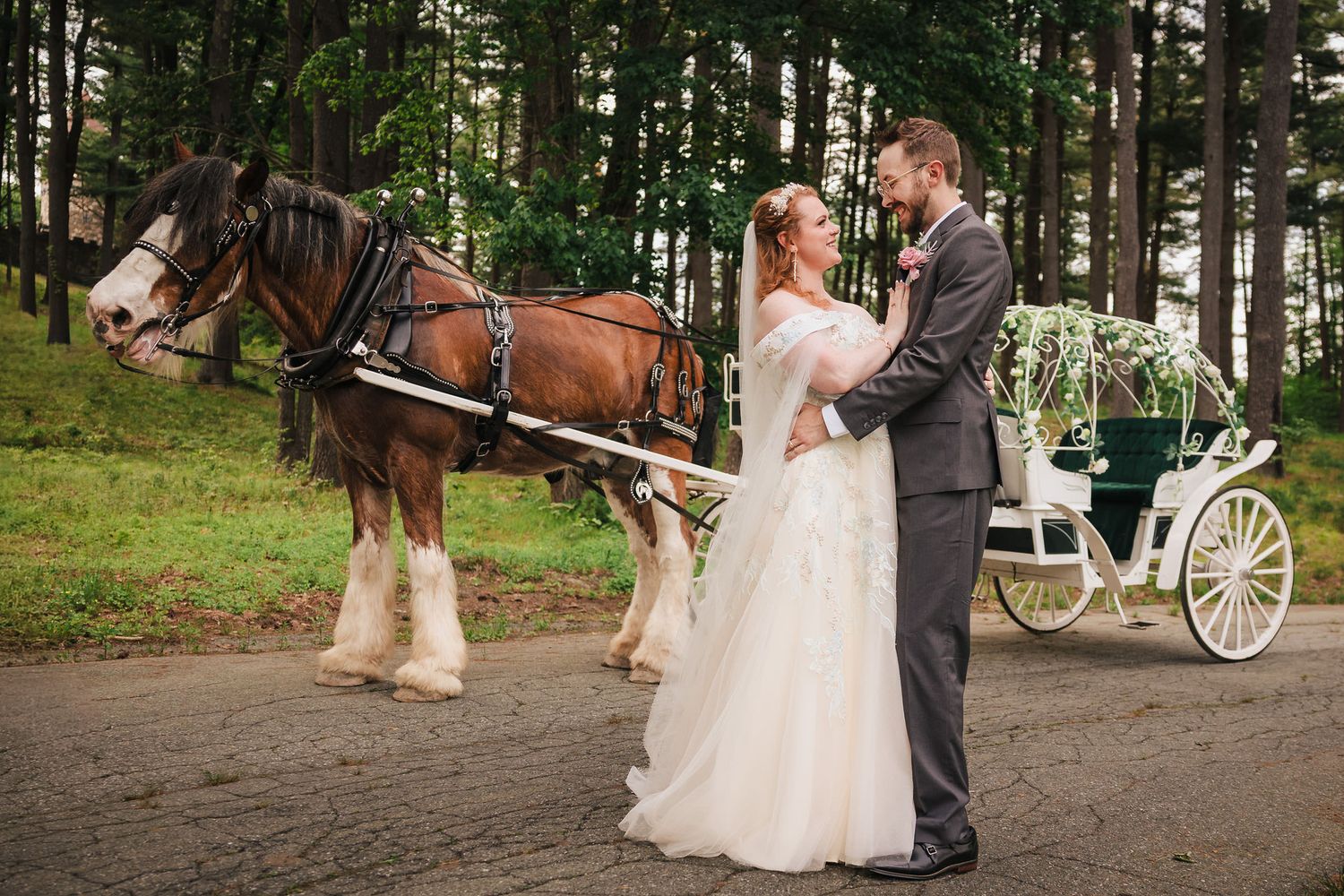 Bride and groom embracing beside a Clydesdale horse, their carriage parked among the trees behind them.