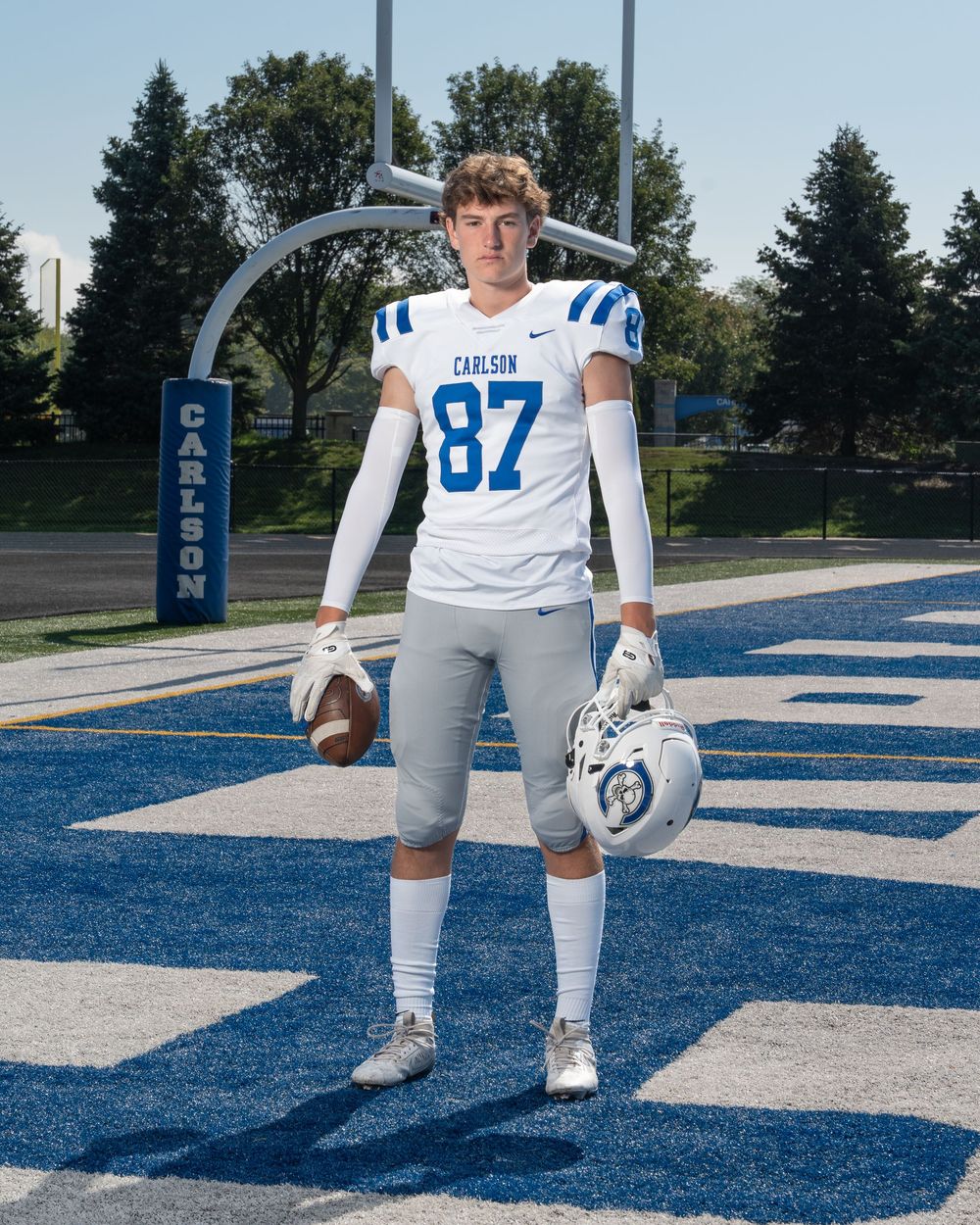 Carlson High School football player #87 poses in white uniform on blue field holding helmet and football.