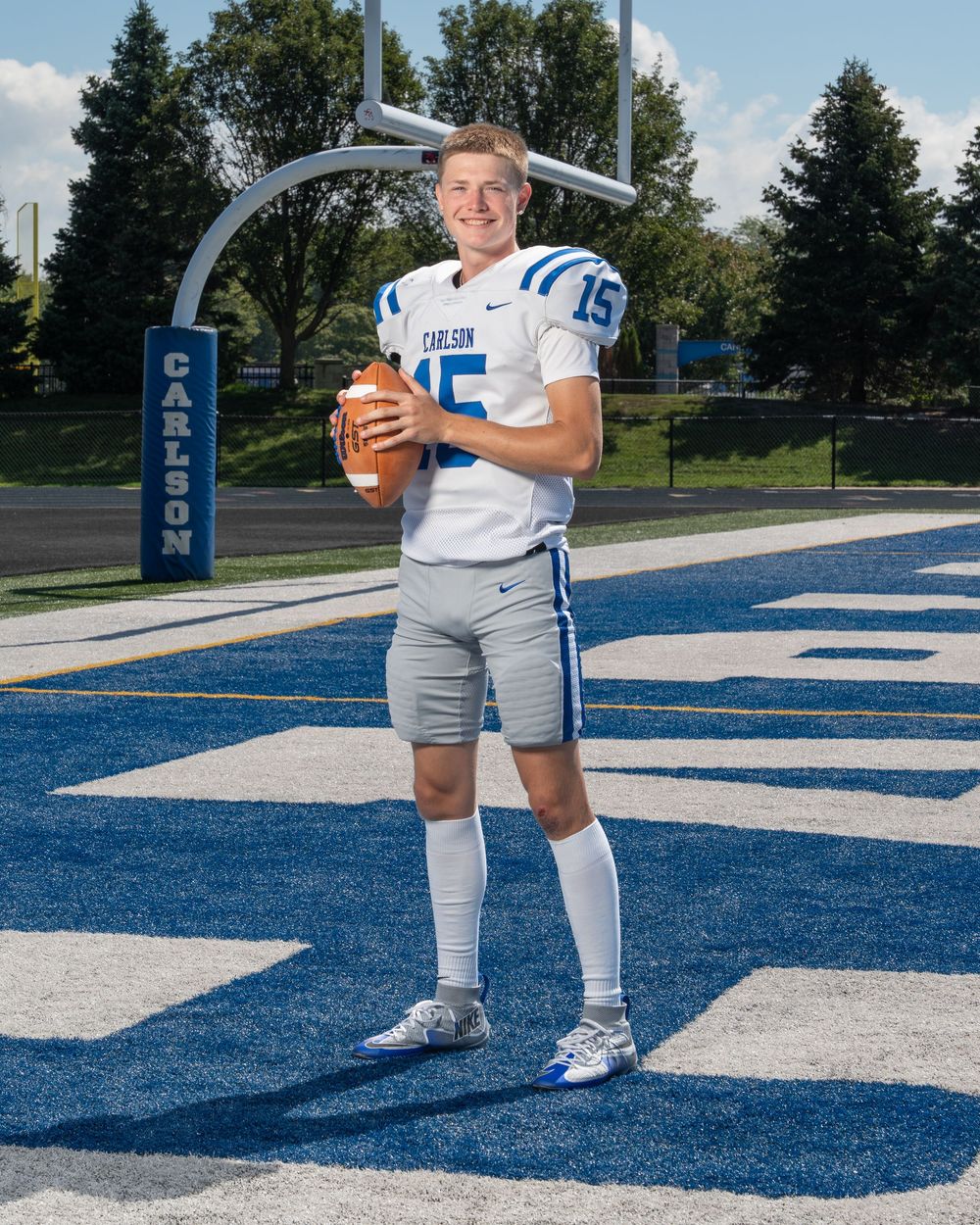 Carlson High School football player #15 poses with a football on a blue turf field in white and blue uniform.