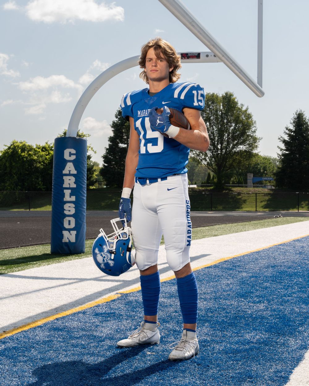 High school football player wearing blue number 15 jersey stands proudly on field holding helmet near Carlson goalpost.