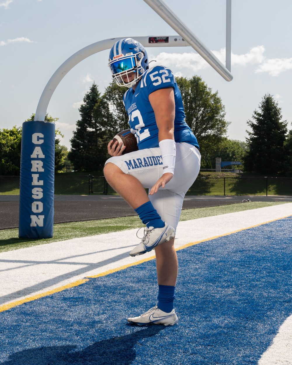 Football player #52 in blue Marauders uniform poses near Carlson goalpost on turf field.