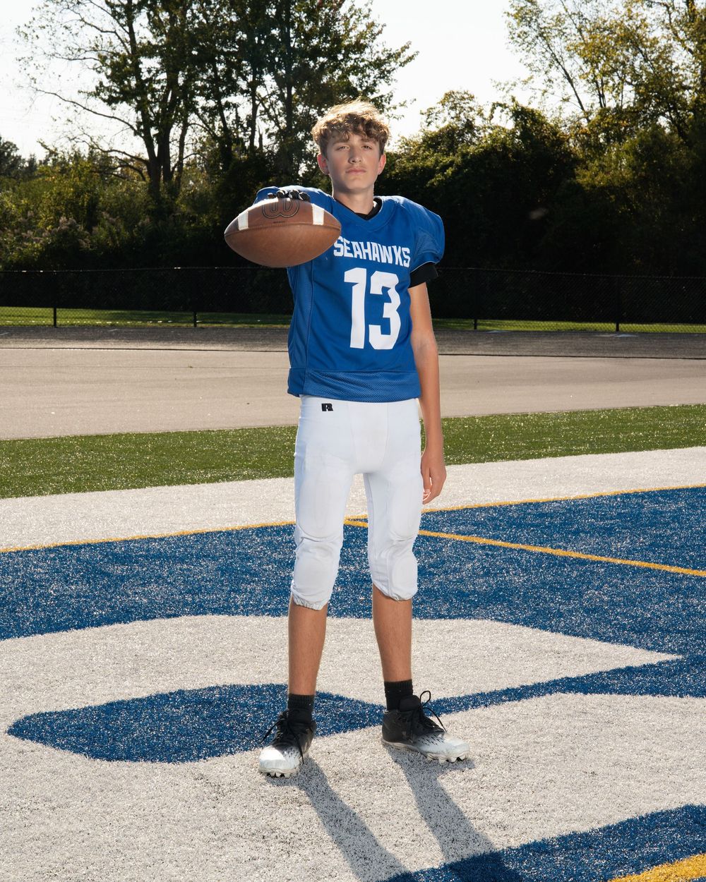 Teen football player #13 in blue Seahawks jersey holds ball on field.