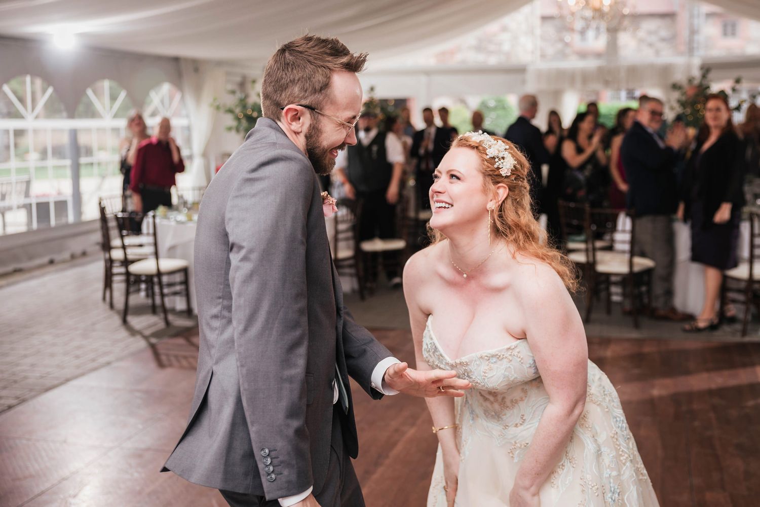 Bride and groom laughing together during their first dance, guests visible seated behind them under the tent.