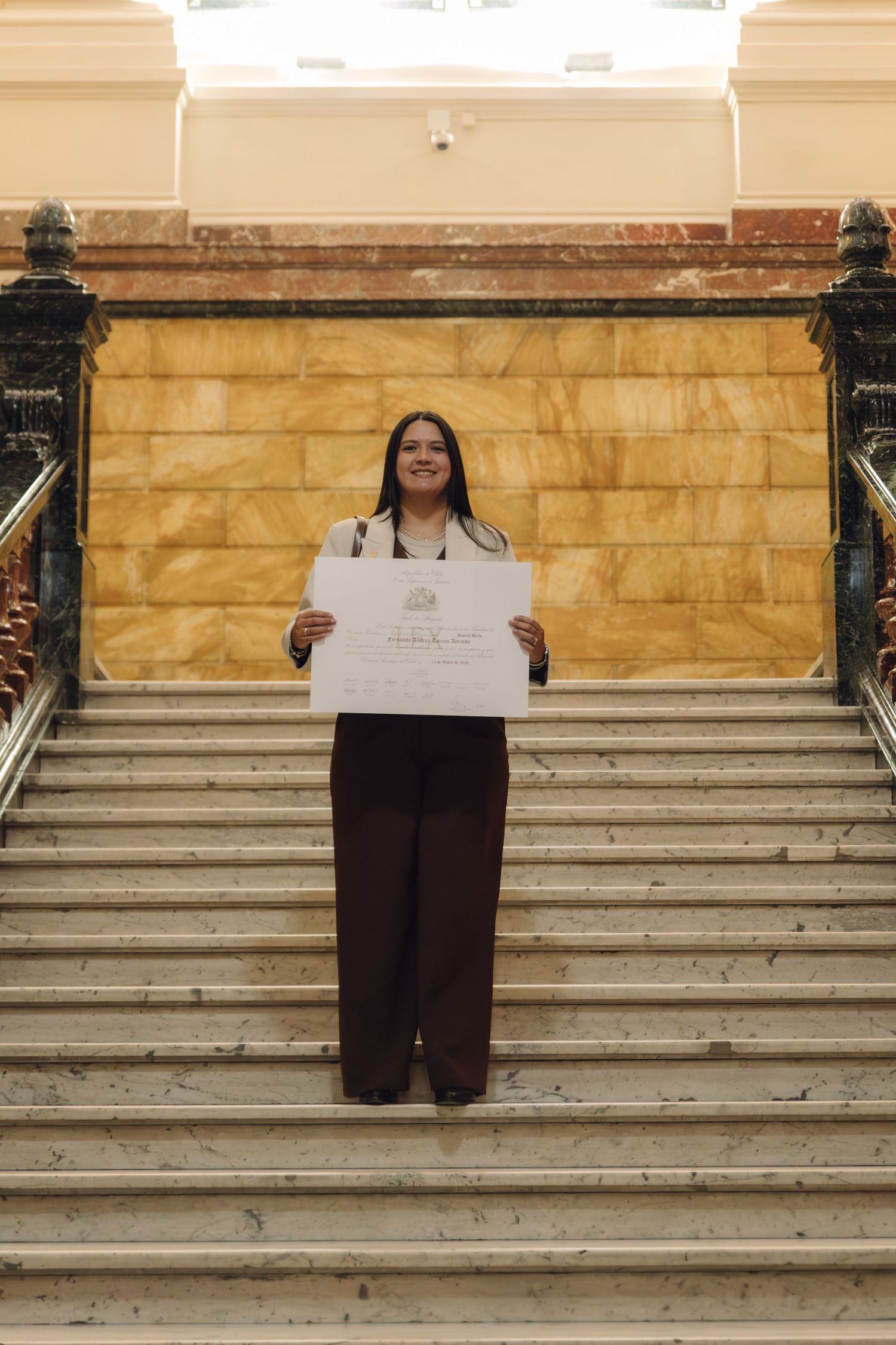 Happy woman in brown wide-leg pants holding a diploma certificate while standing on grand marble stairs in a formal building.