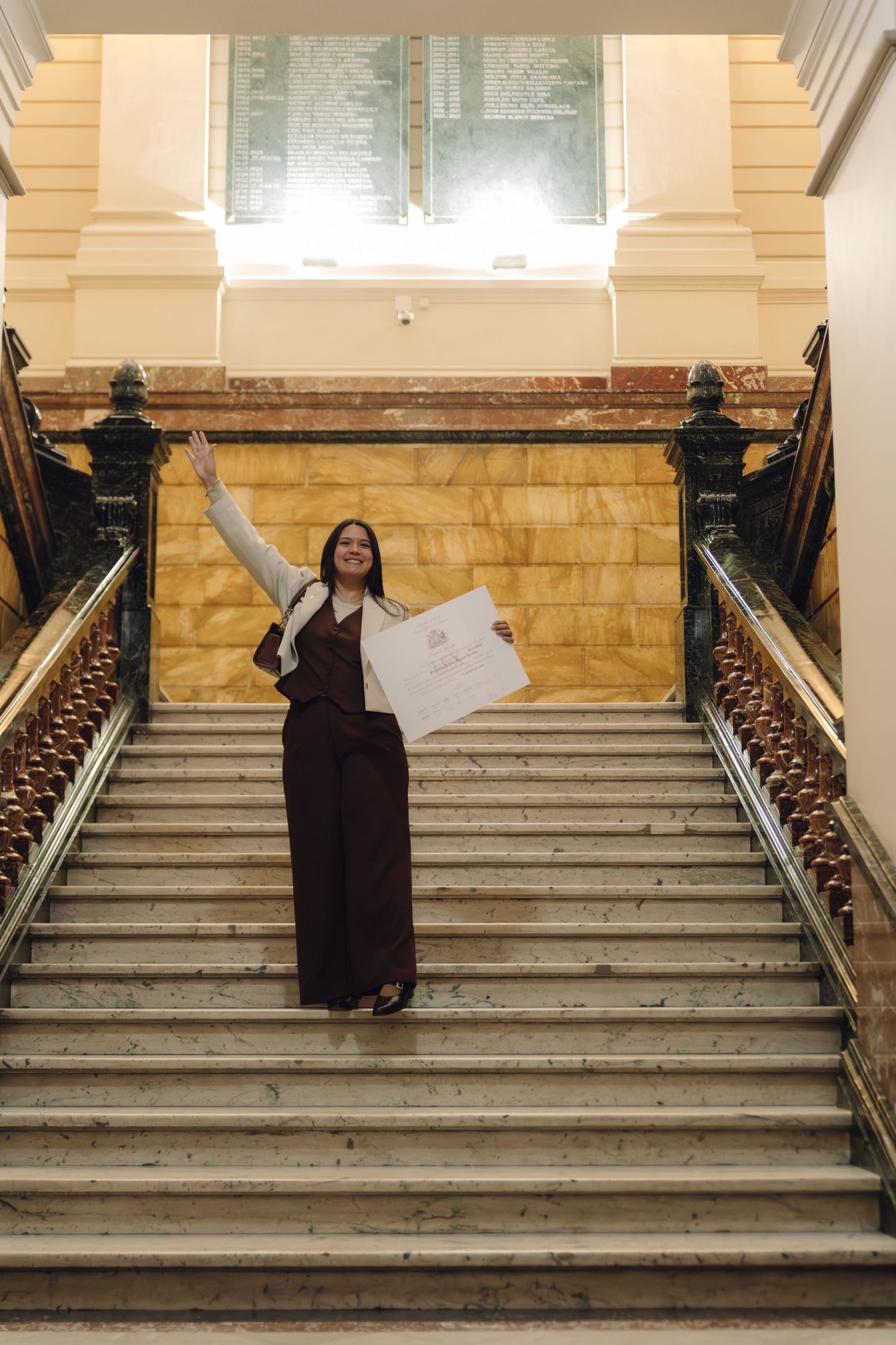 Joyful woman celebrating on grand marble staircase, raising one arm and holding a diploma certificate in an elegant building.