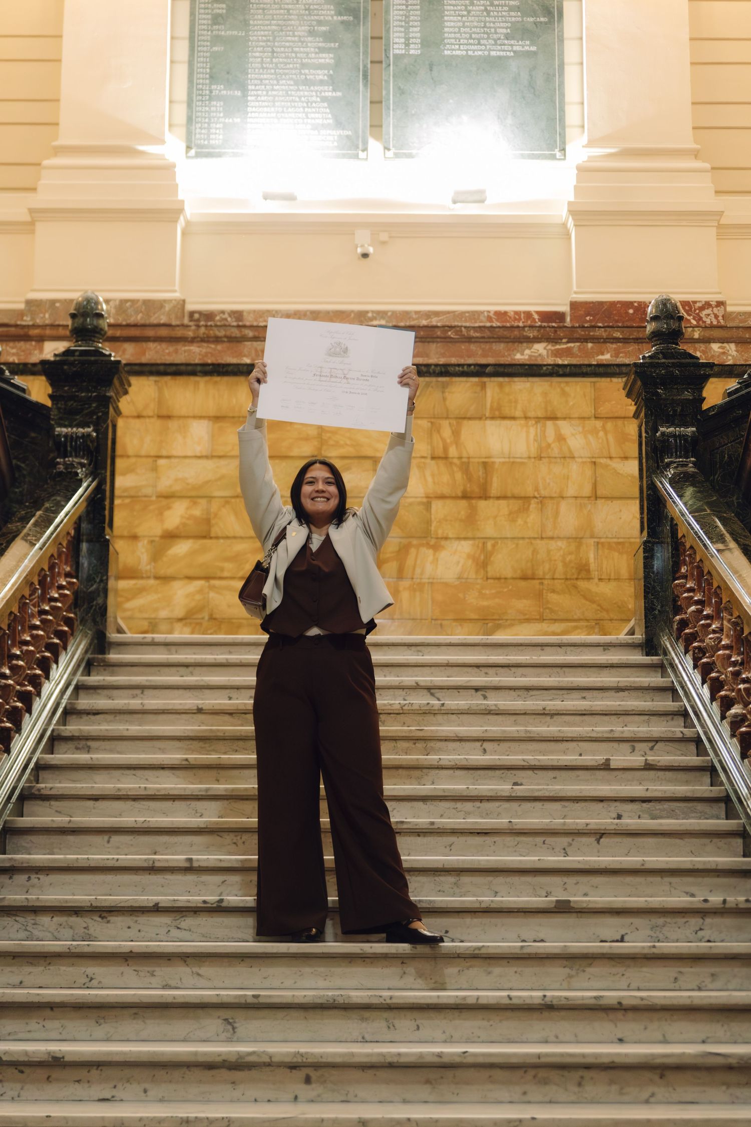 Proud woman holding diploma certificate above her head while standing on grand marble staircase in a formal ornate building.