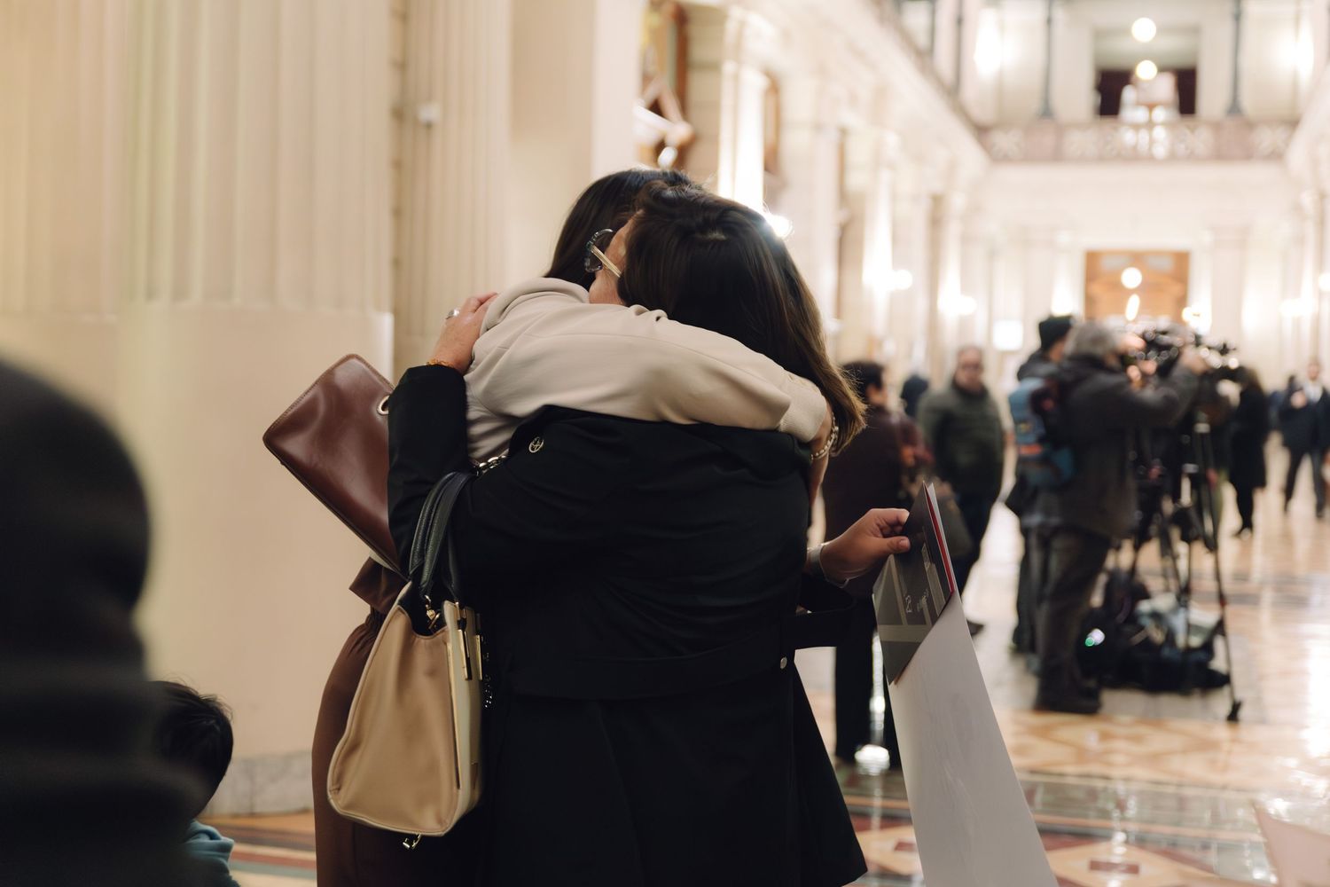 Two women sharing a warm embrace in an elegant columned hallway after a graduation ceremony.