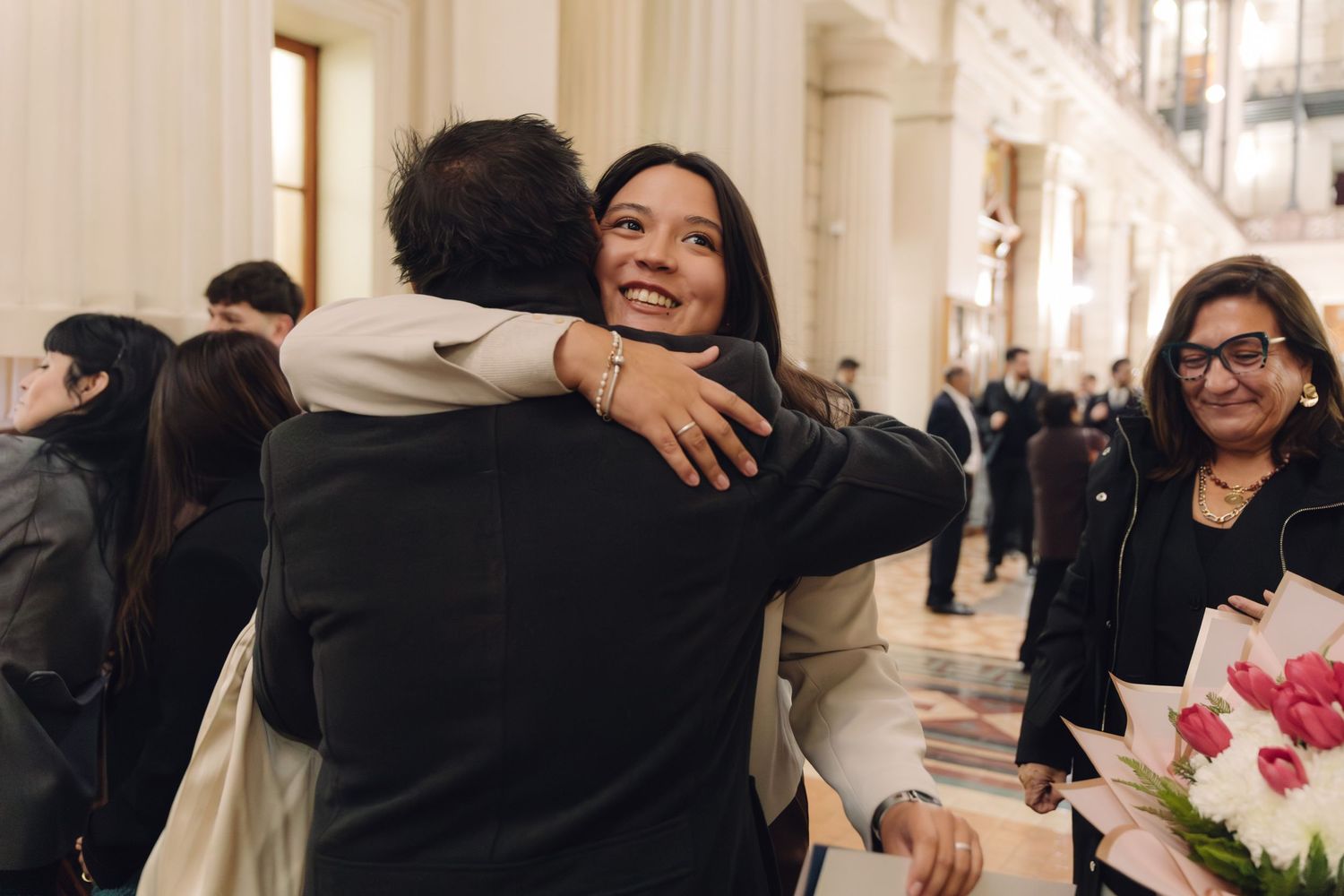 Smiling graduate hugging someone while holding pink tulips, with guests celebrating in an elegant ceremonial hall.