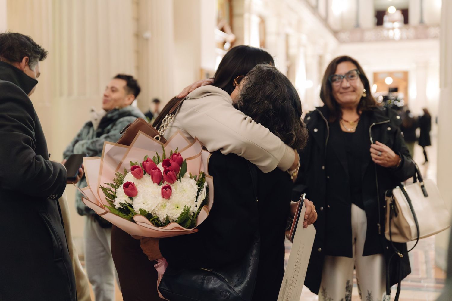 Two people share a warm hug while one holds a bouquet of red and white flowers in an elegant hall.