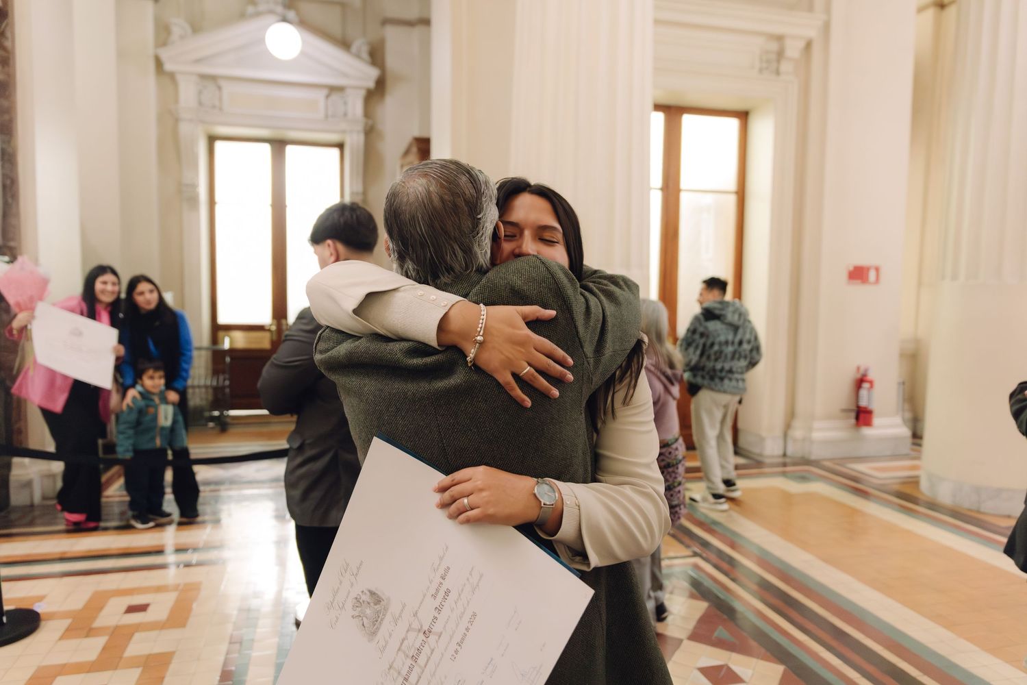 Two people embrace warmly in a grand hall, one holding official documents after a graduation ceremony.