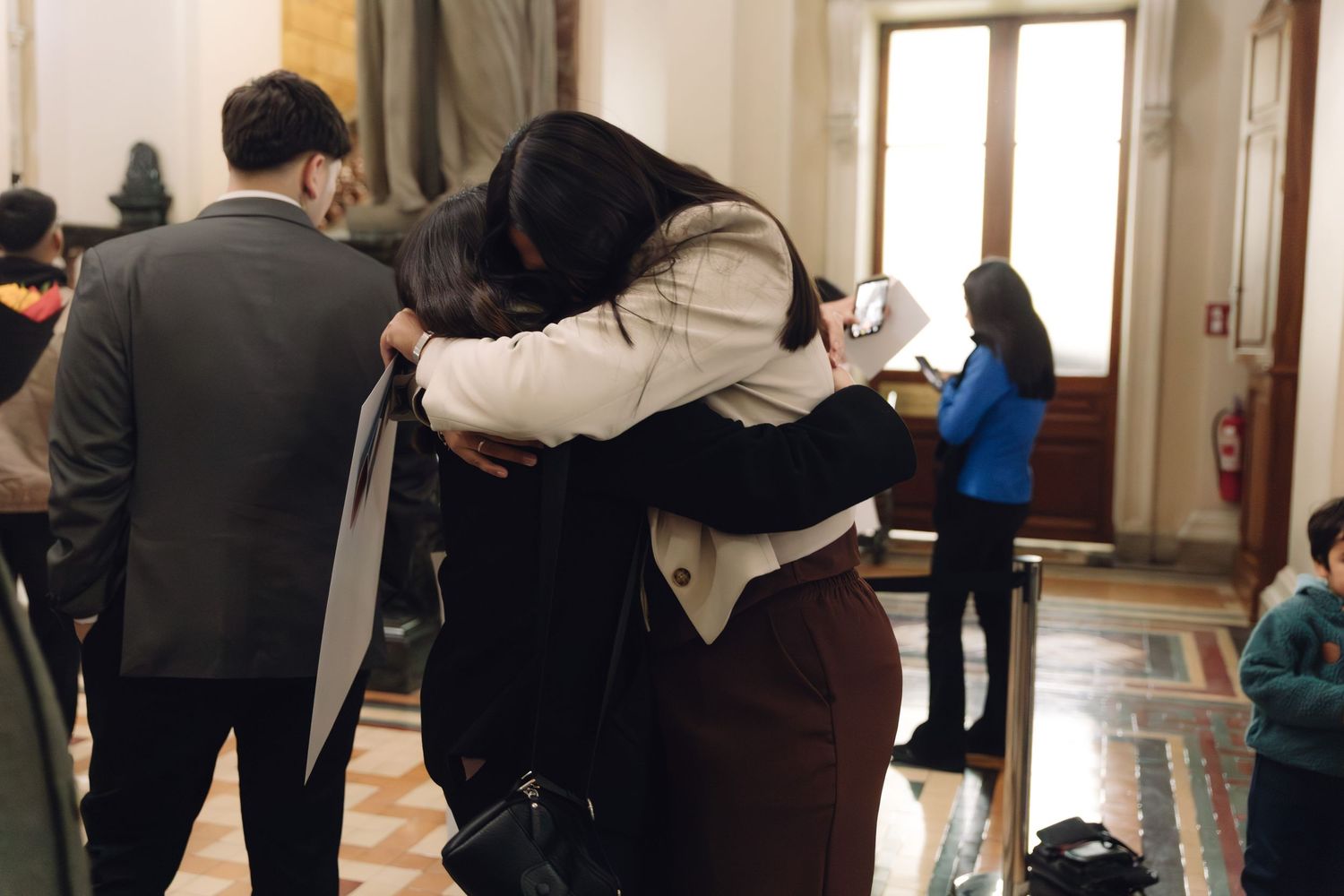 Two graduates share a heartfelt hug in an ornate marble-floored hallway during a graduation celebration.
