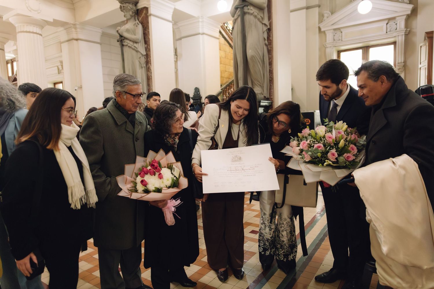 Group of people celebrating a graduation, holding flowers and official certificates in an elegant venue.