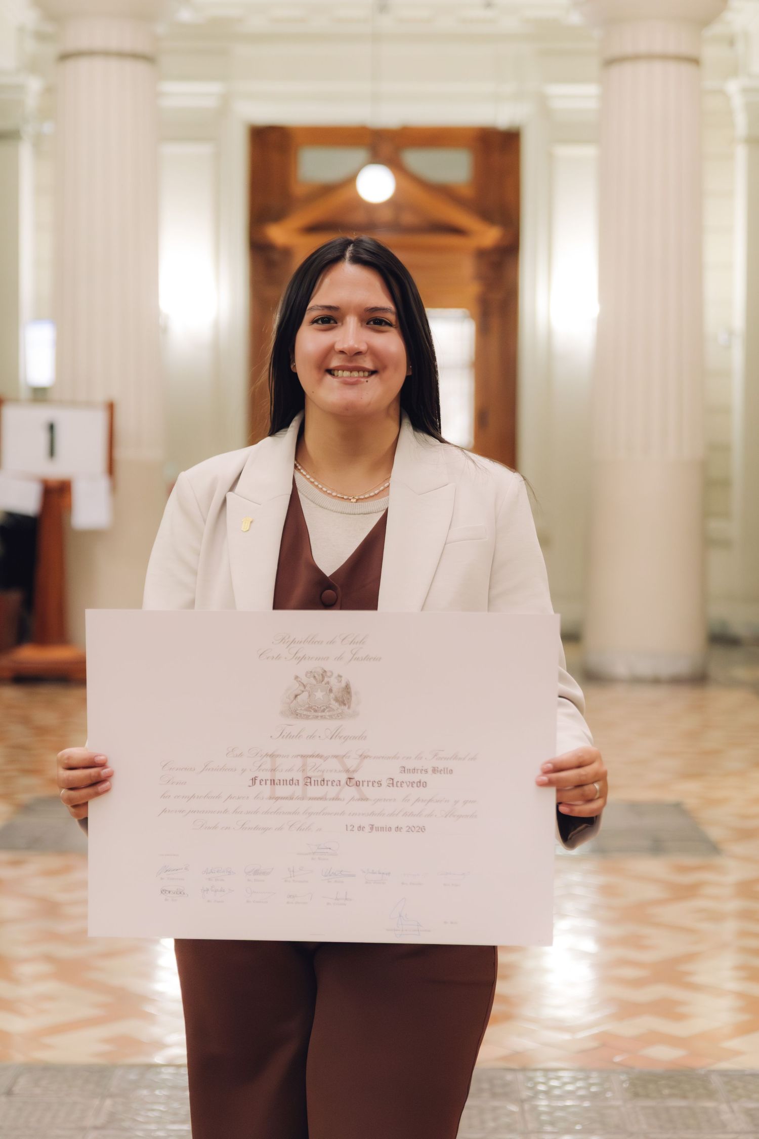 Smiling young woman in white blazer and brown suit proudly holds her graduation diploma in a grand hall.