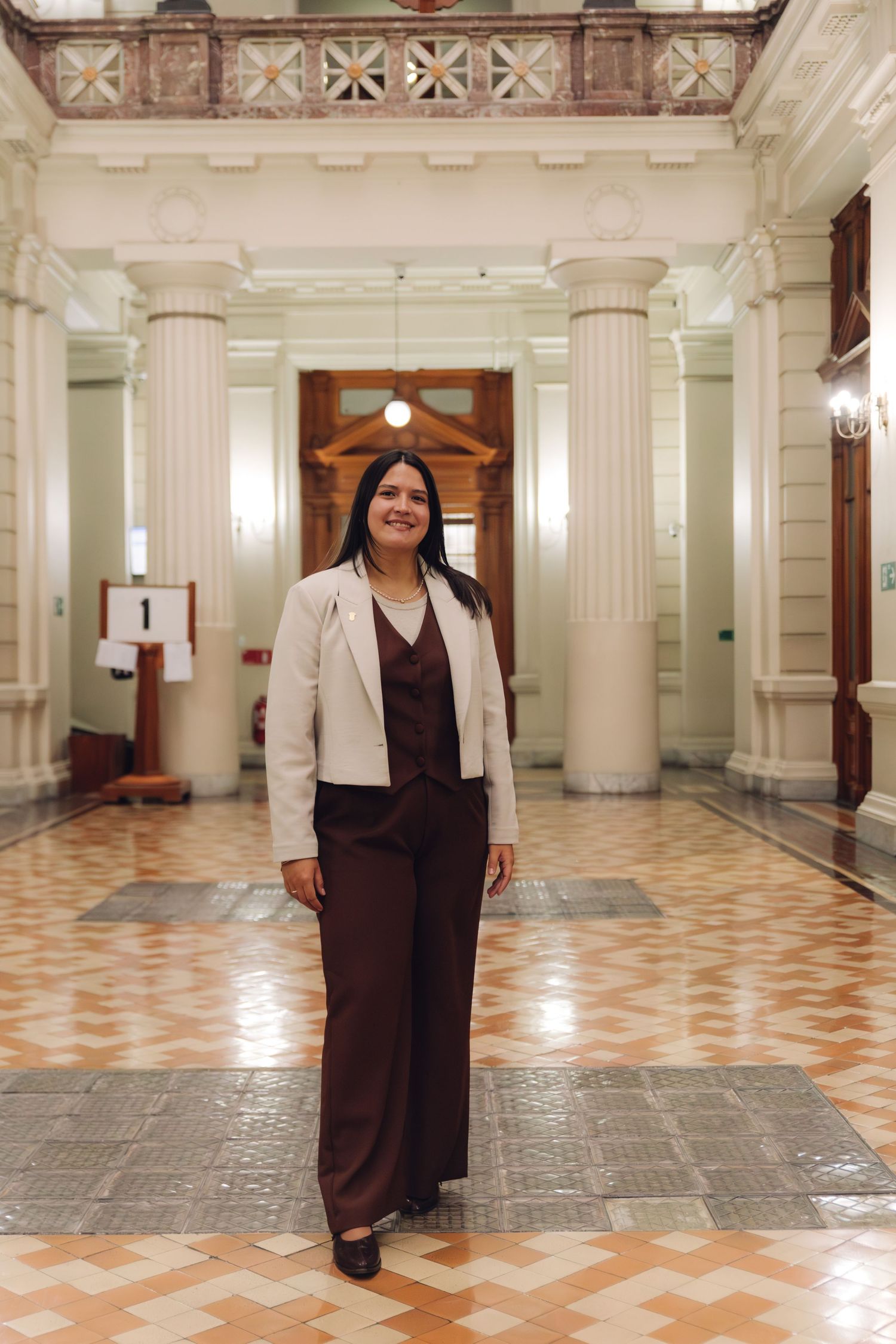 Confident young woman in white blazer and brown pantsuit poses in an elegant columned hallway.