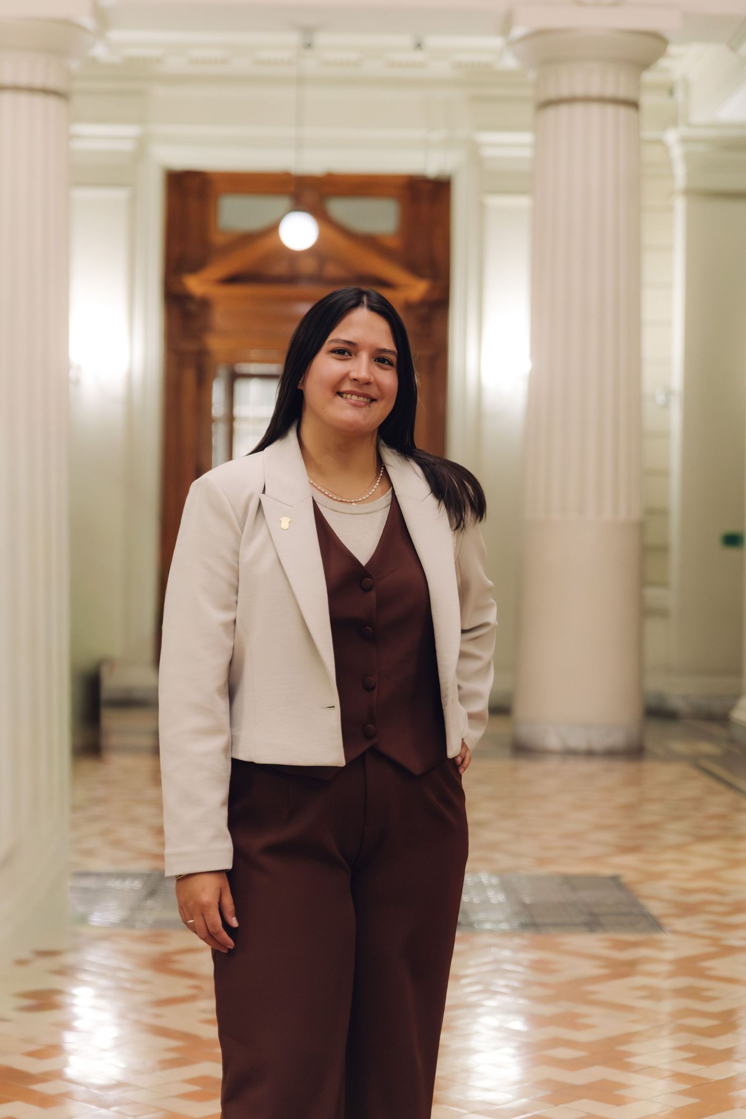 Young woman in white blazer and brown suit smiles confidently in an elegant columned corridor.