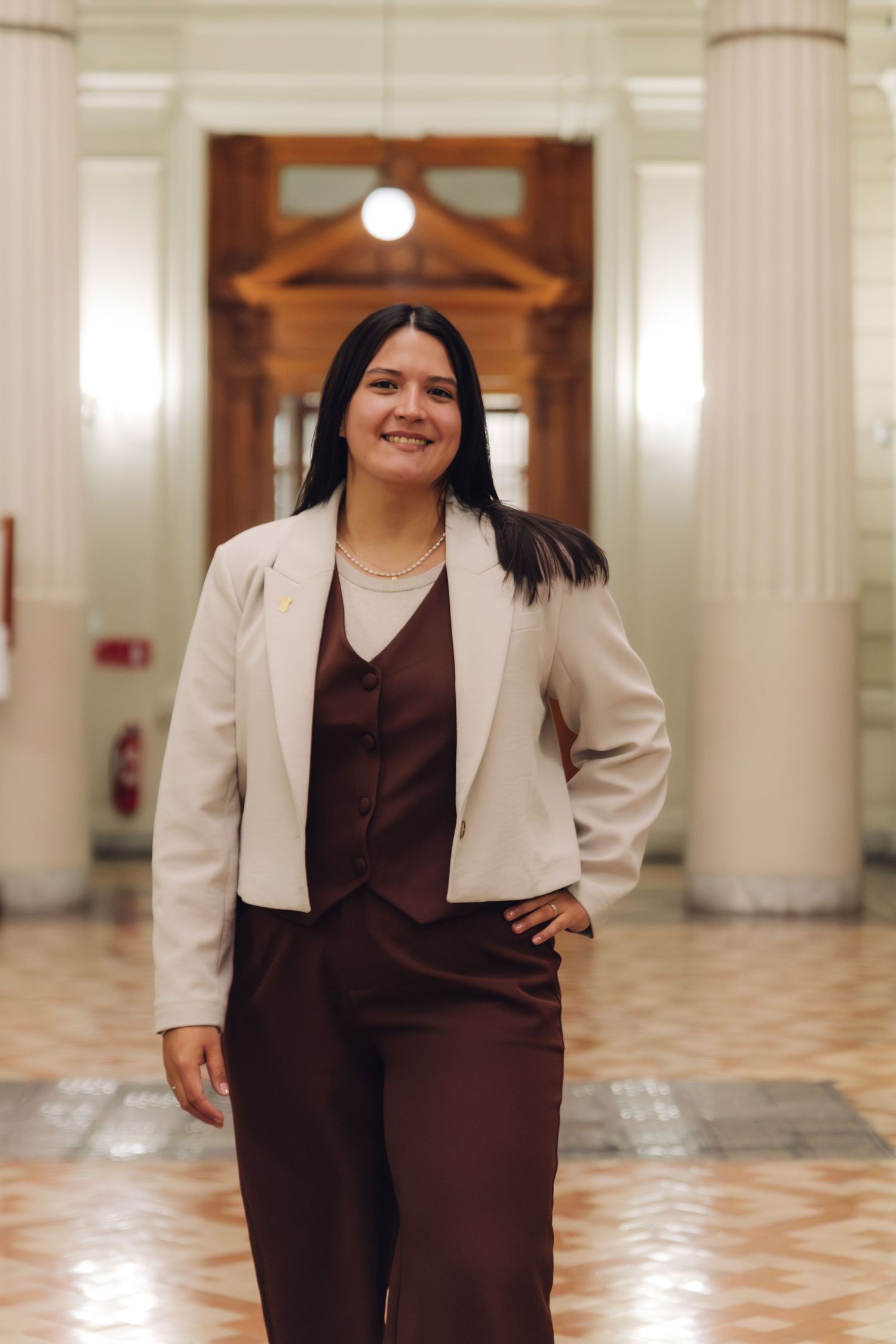 Smiling young professional woman in white blazer and brown suit poses in a grand classical hallway.