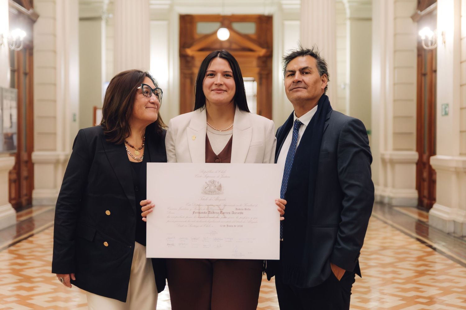 Three people smile together in a grand corridor while displaying an official framed certificate.