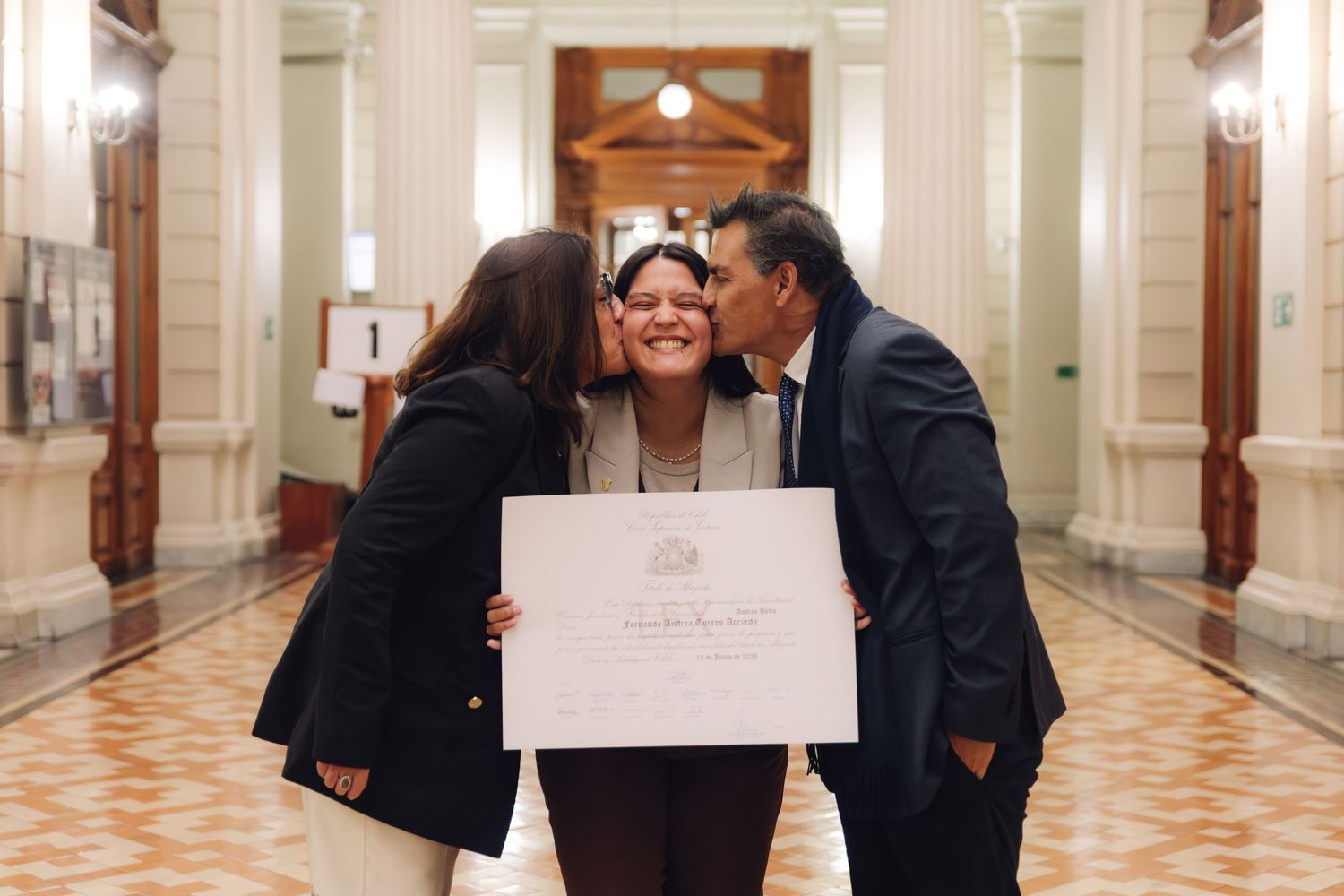 Two people kiss the cheeks of a certificate holder in a celebration photo in a formal hallway.