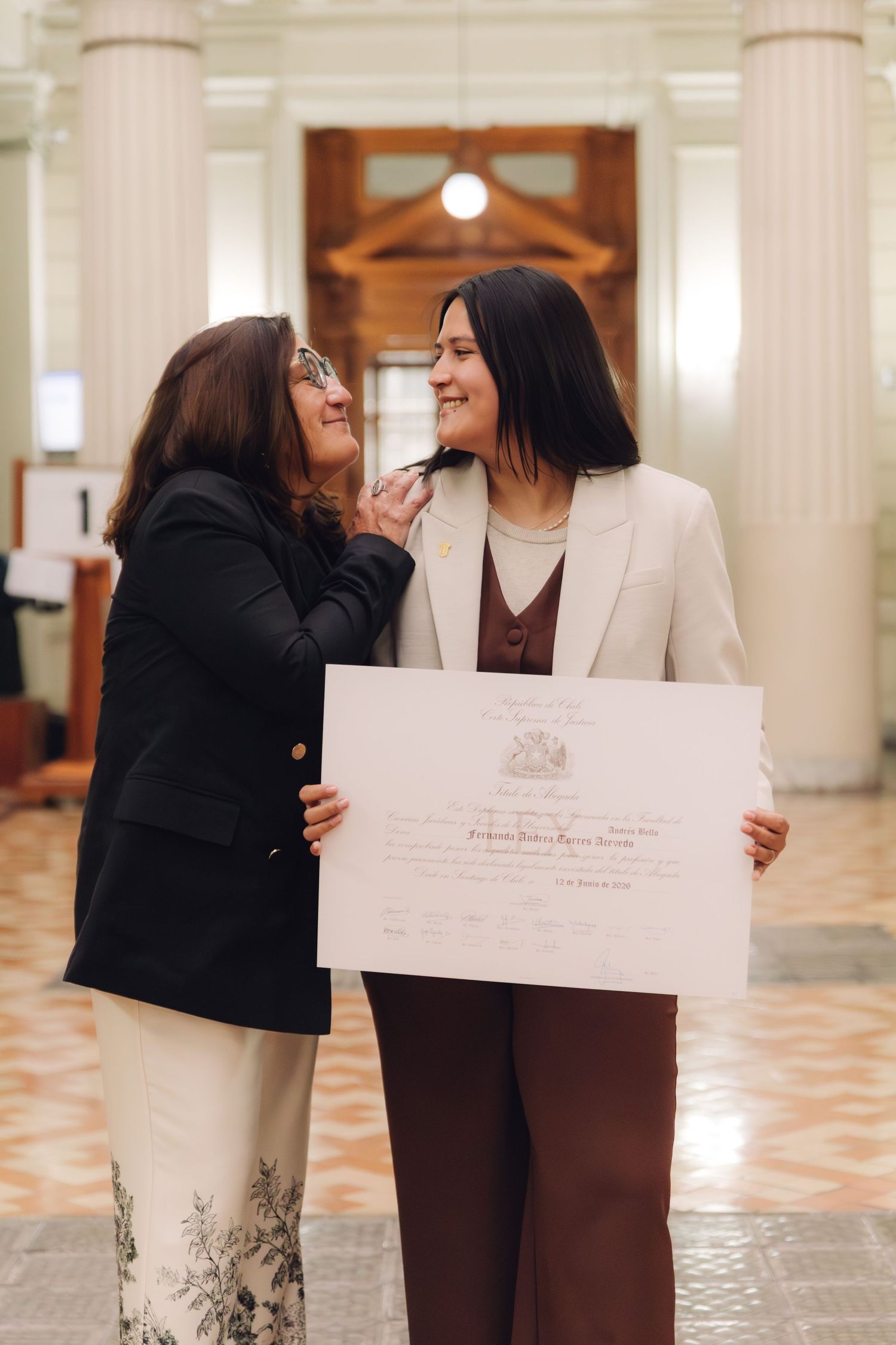 Two women share a warm moment holding an official certificate in an elegant columned building.