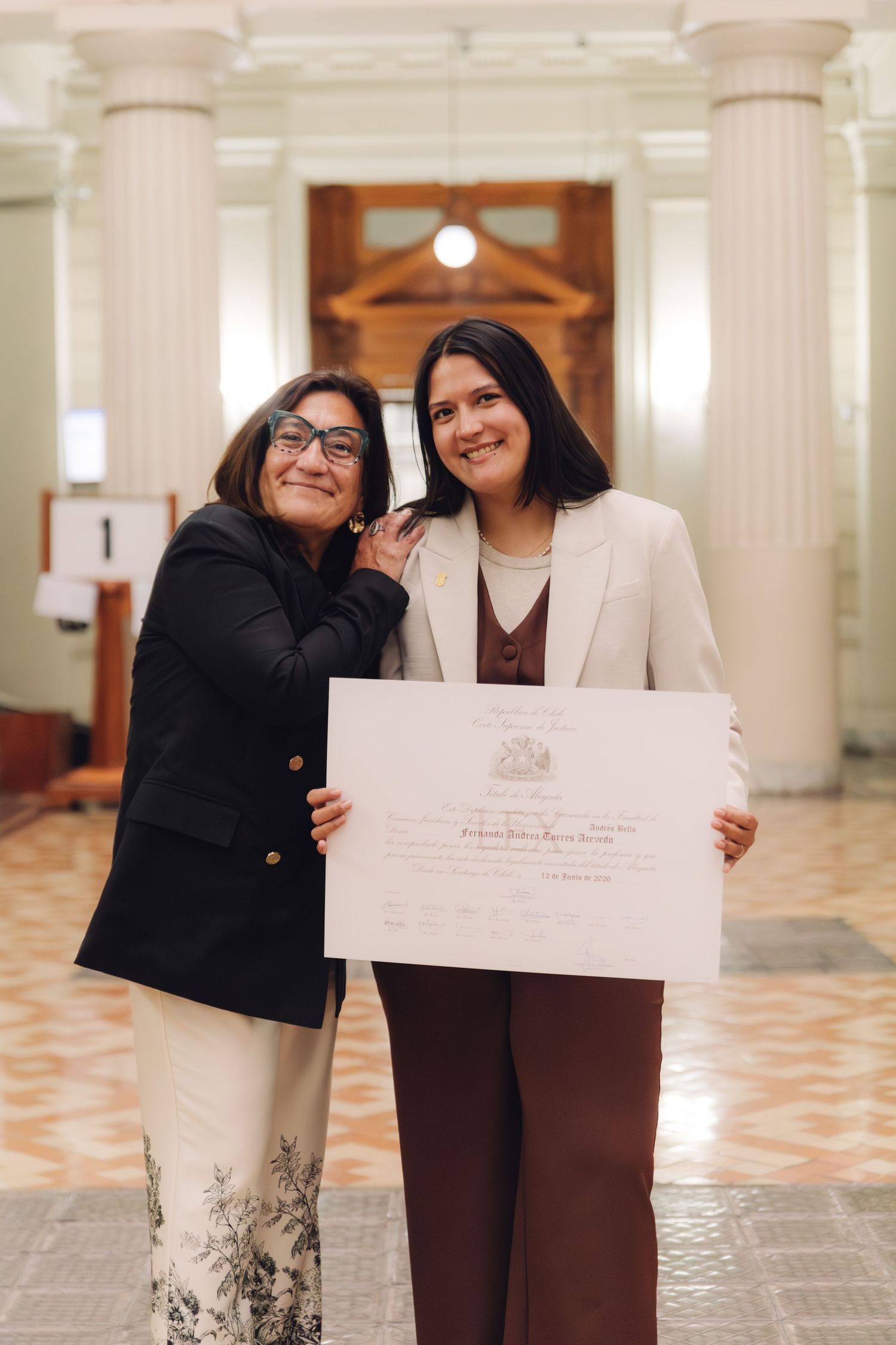 Two smiling women pose together holding a large official certificate in a grand interior hallway.