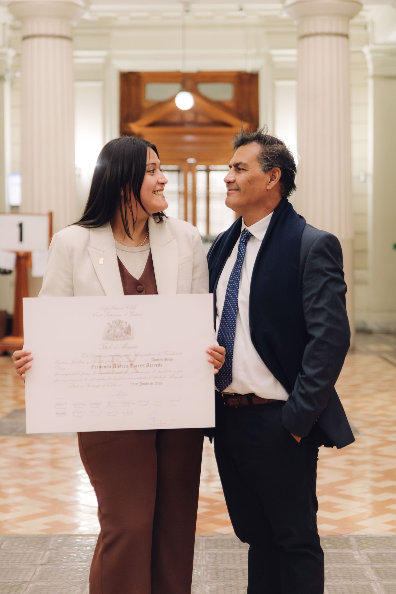 A young woman and an older man share a proud moment holding an official certificate indoors.