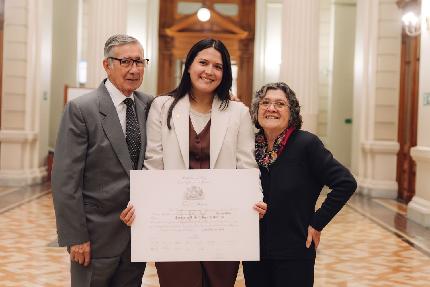 Three people pose together proudly displaying a large official certificate in an elegant corridor.