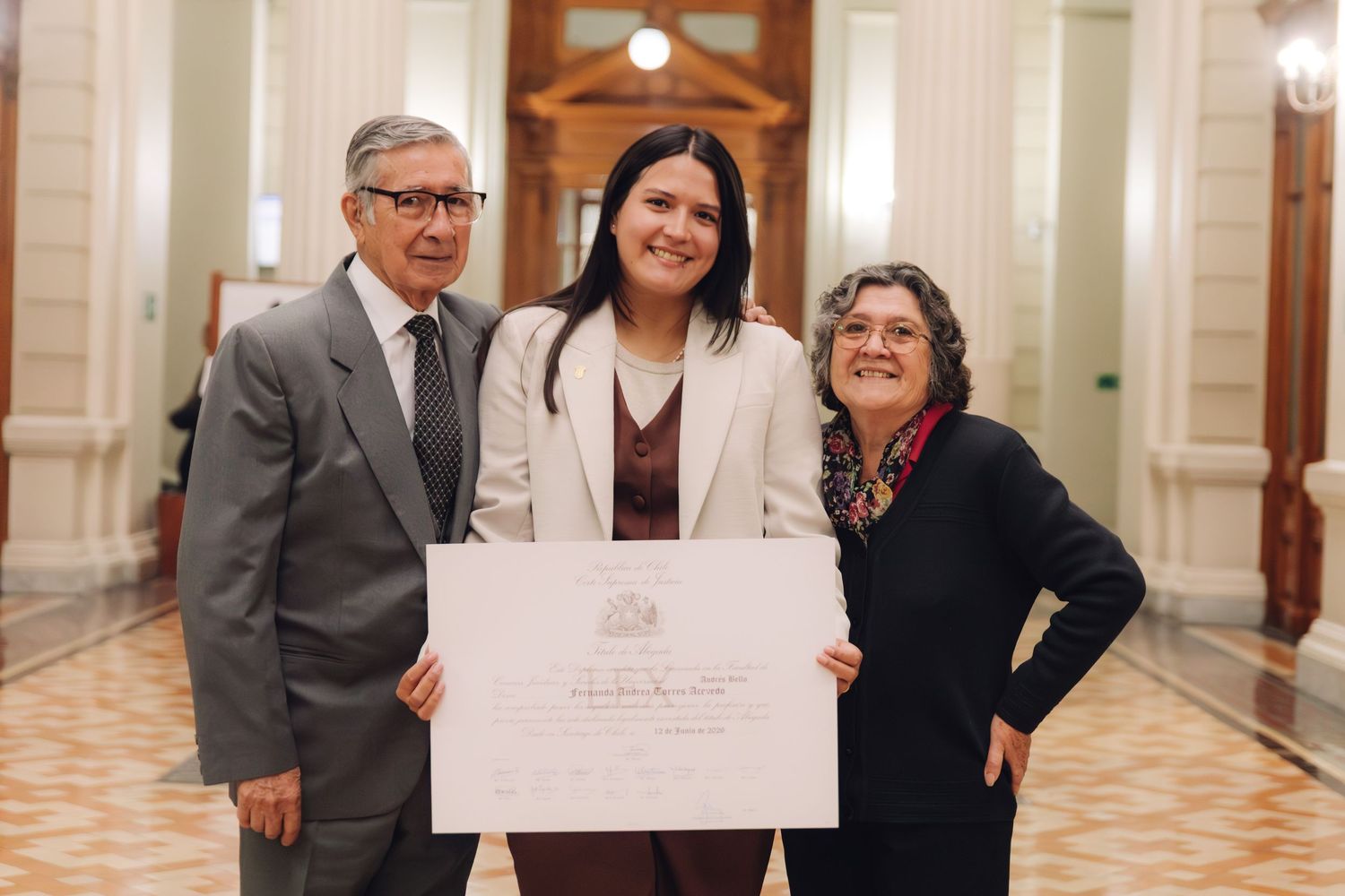 Three people stand together smiling while holding a large official certificate in a formal hallway.