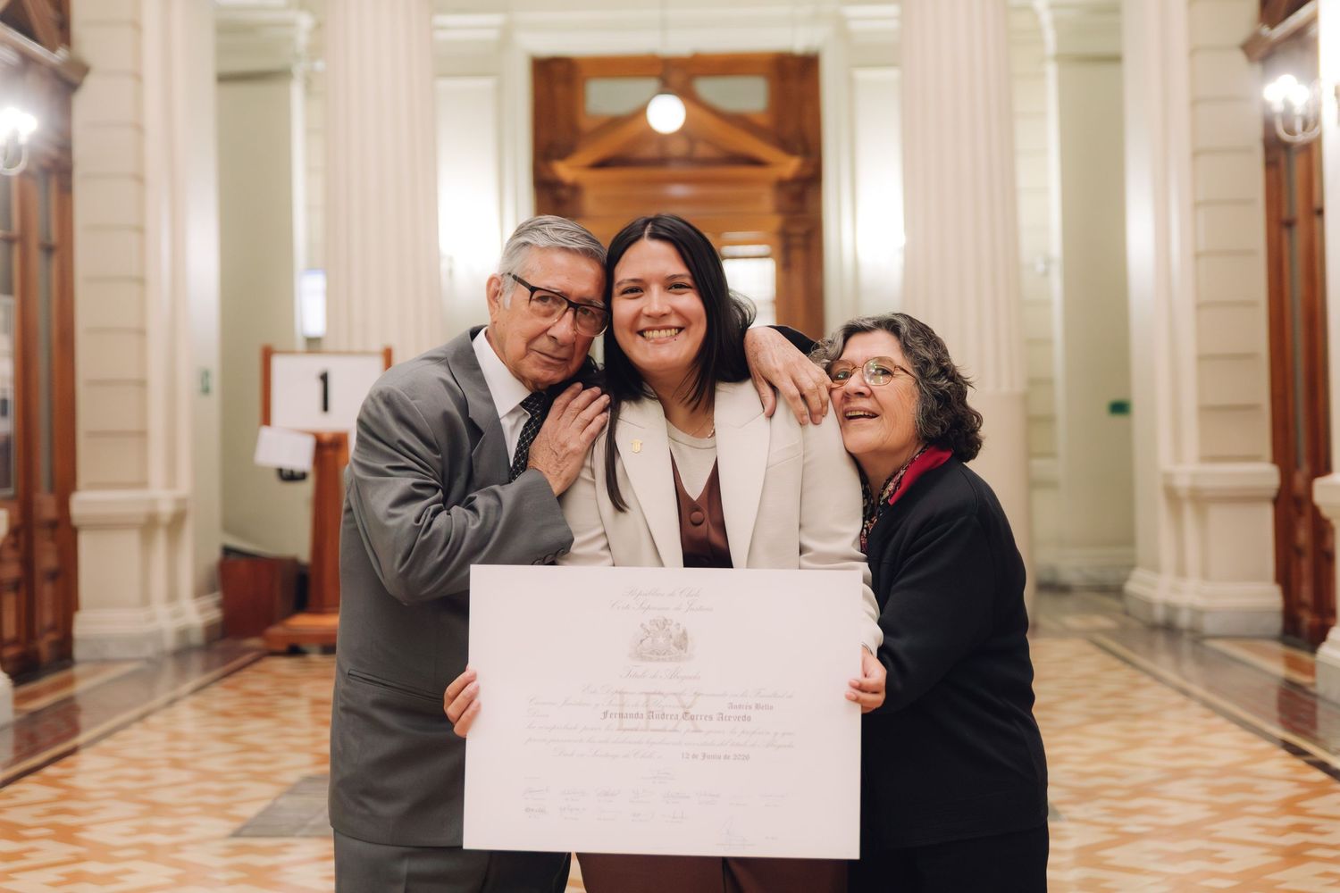 Three people celebrate joyfully in a grand hallway, holding an official certificate or diploma.