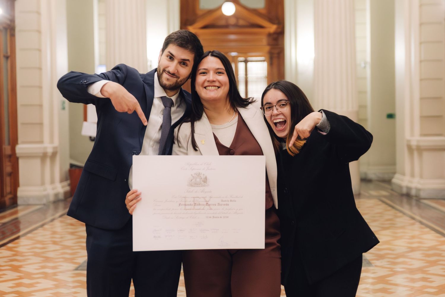 Three friends celebrate joyfully pointing at graduation diploma in ornate hallway.