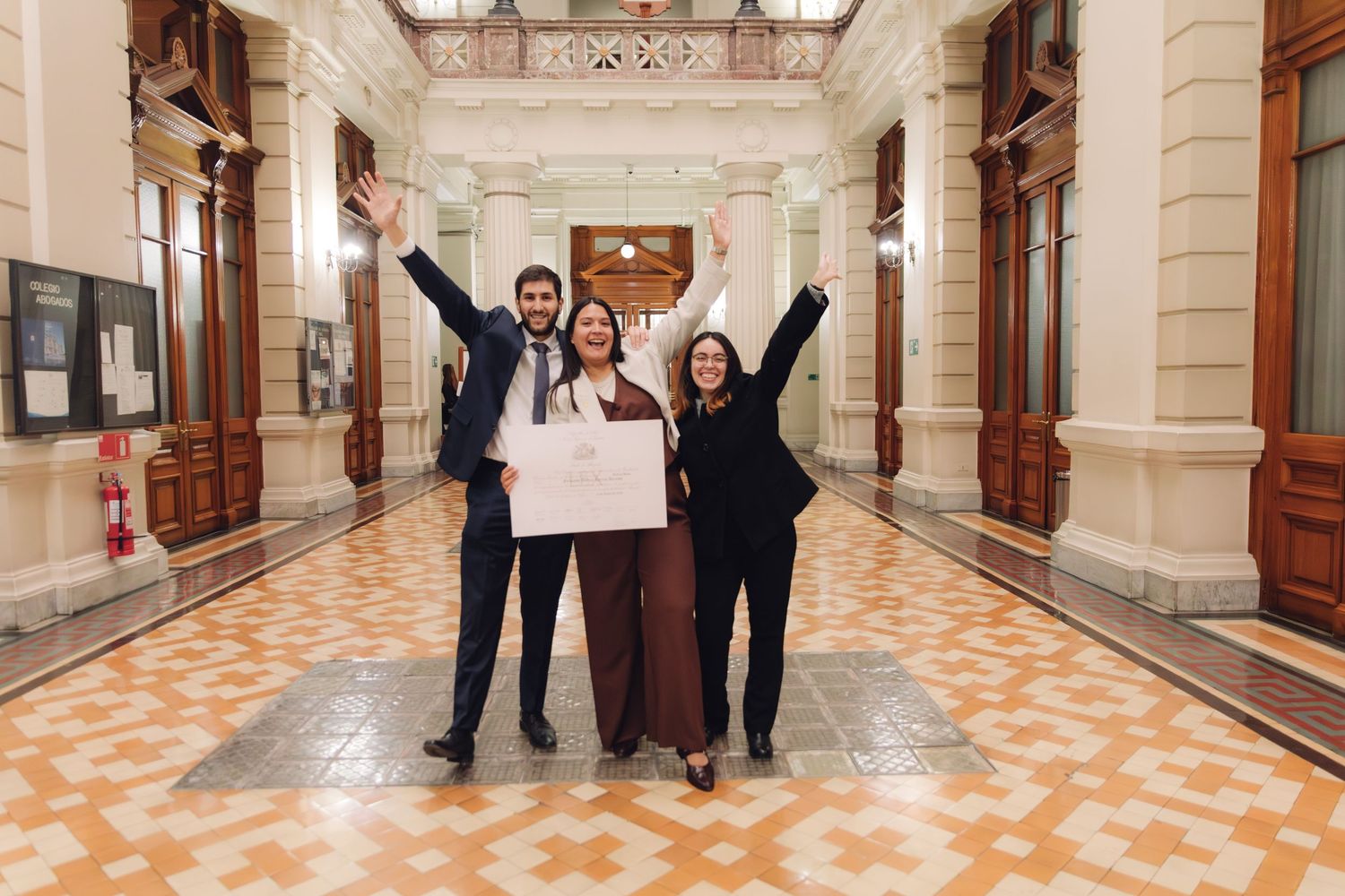 Three graduates celebrate with raised arms holding diploma in classical building.