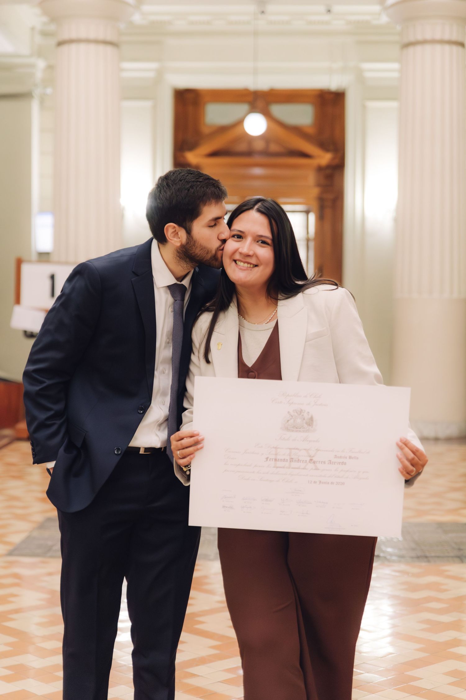 Man kisses woman on cheek as she holds graduation diploma in hall.