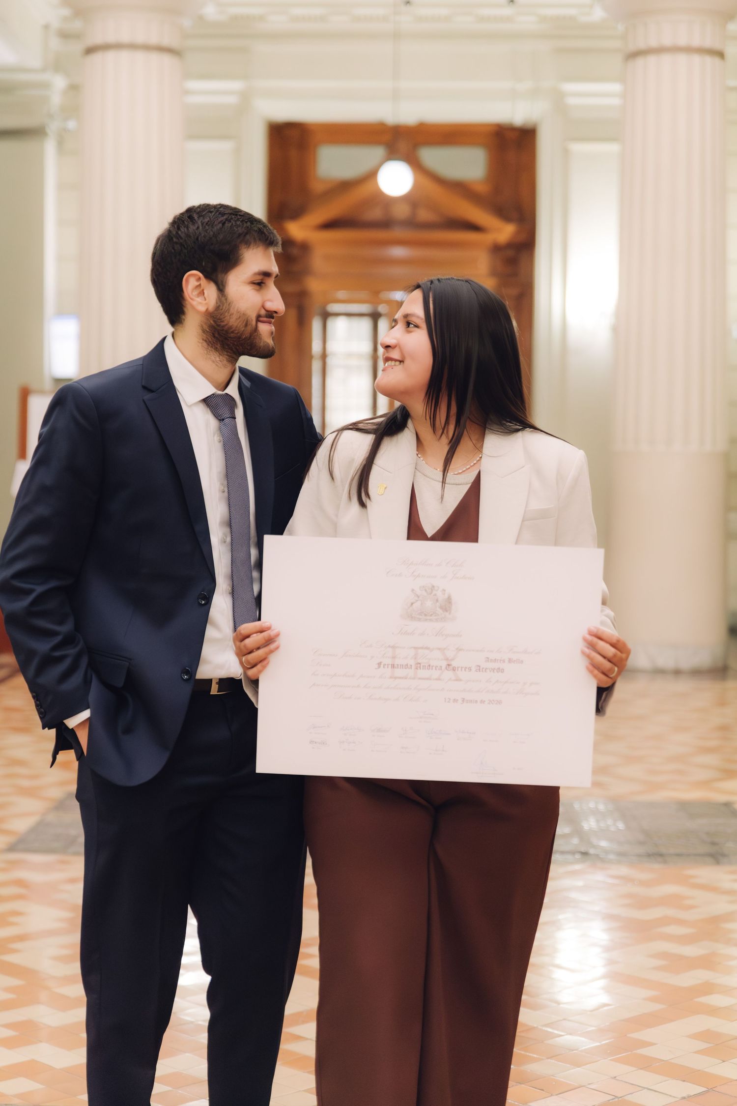 Couple smiles at each other while holding official graduation certificate indoors.