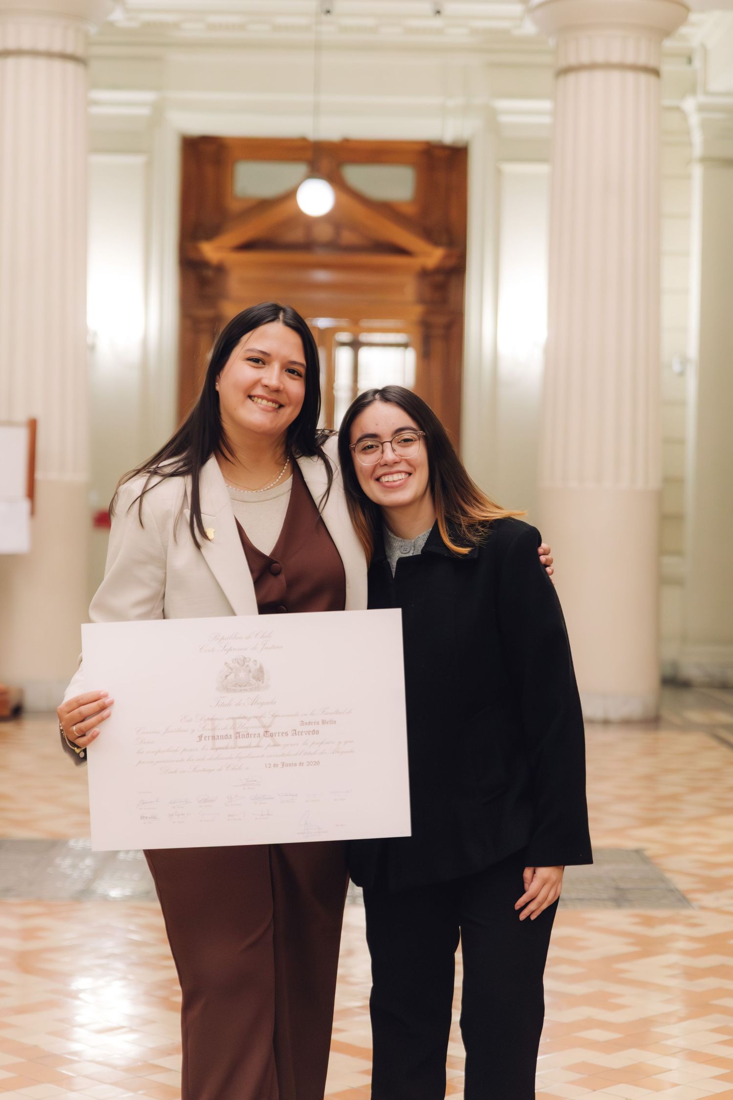 Two young women smile together holding a graduation diploma in formal hall.