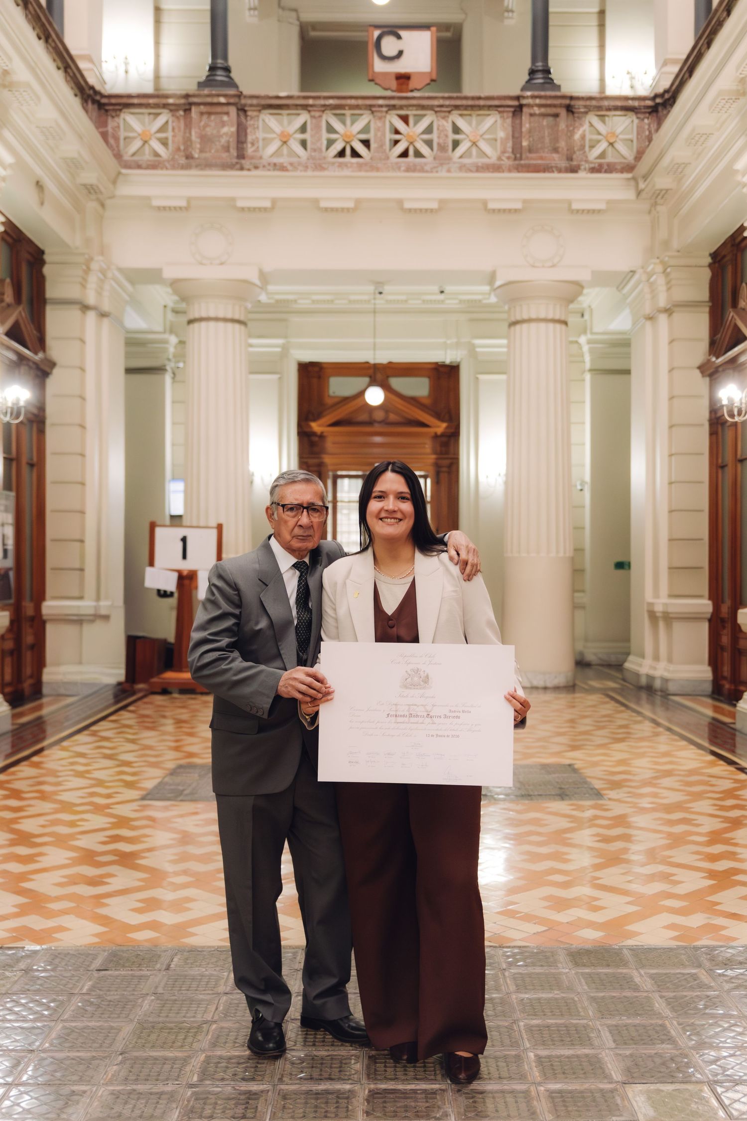 Older man and young woman pose with graduation diploma in grand hall.