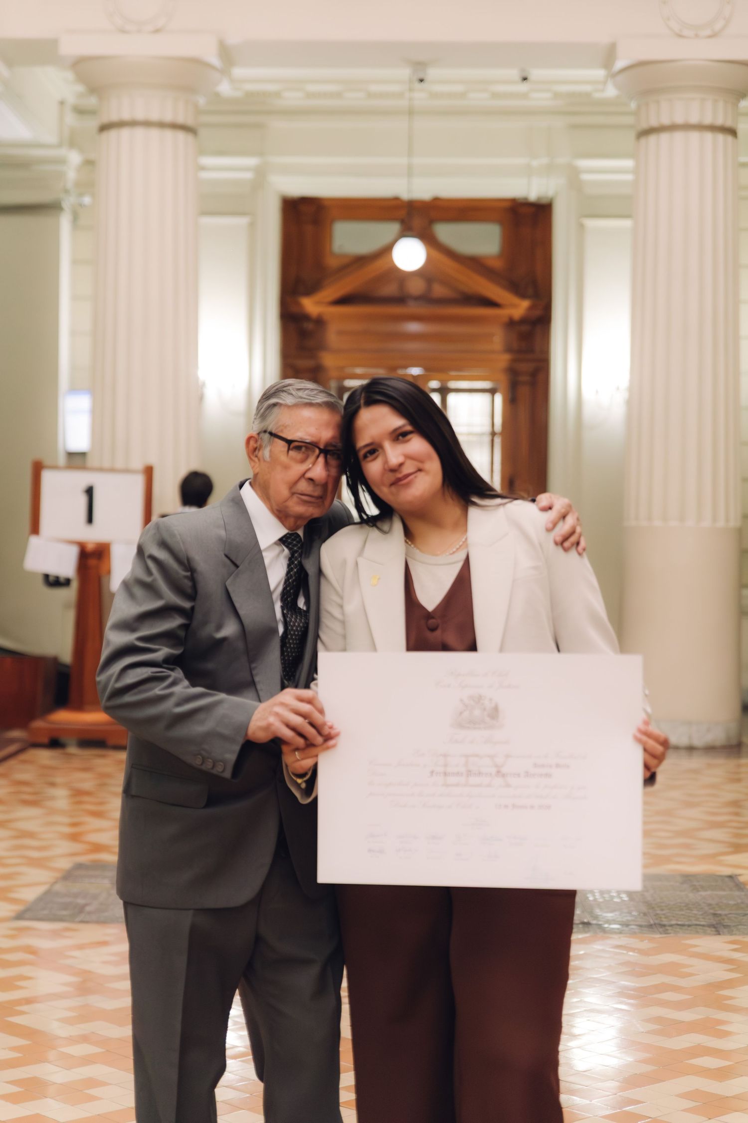 Senior man embraces young woman holding graduation certificate in classical building.