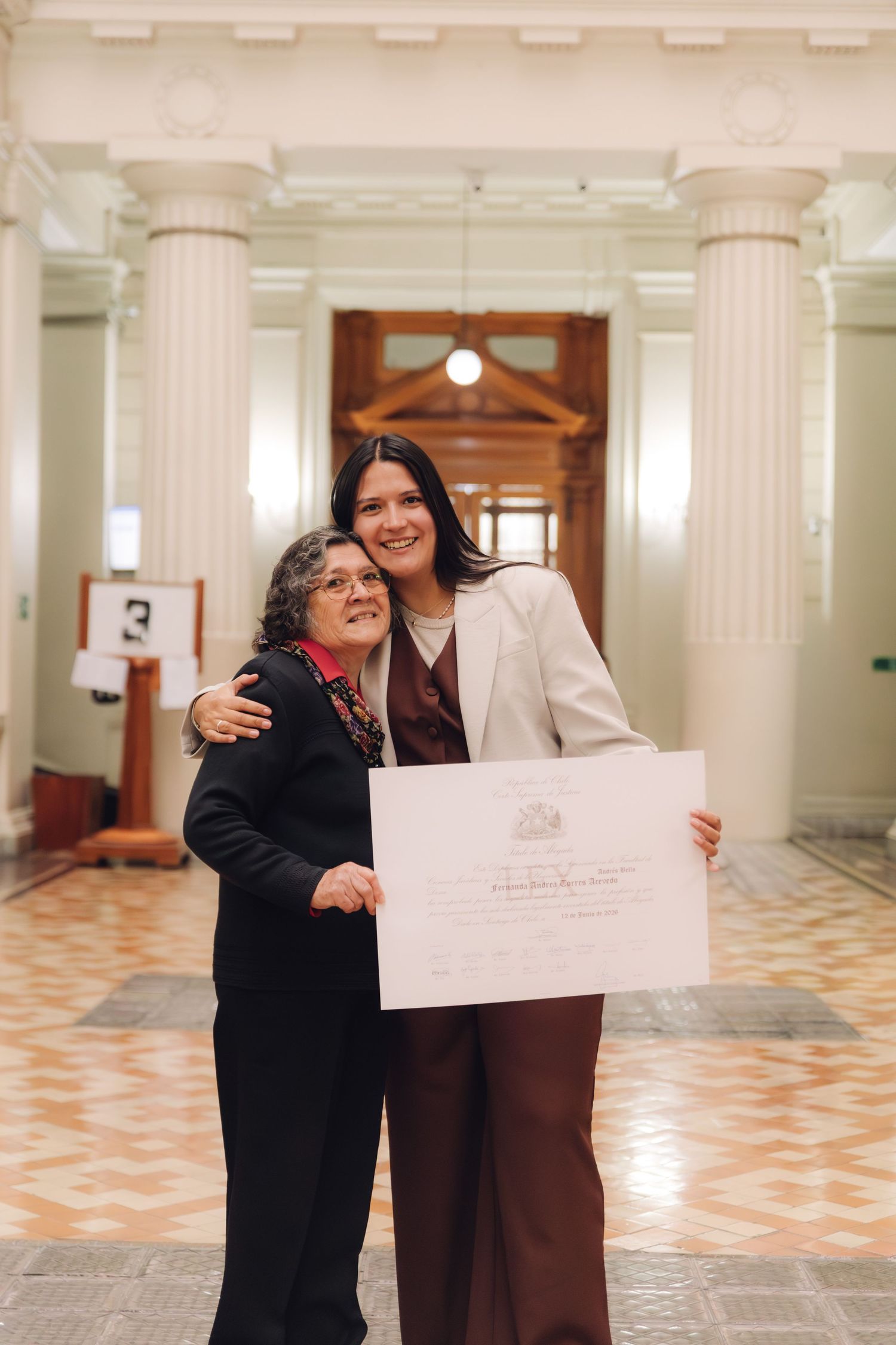 Older woman hugs tall graduate holding diploma in ornate columned hall.