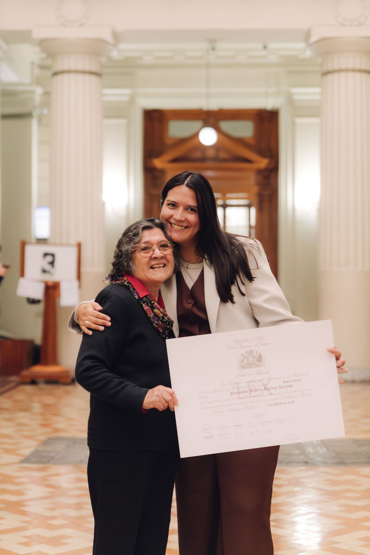 Woman in white blazer holds certificate with elderly woman in formal hall with columns.