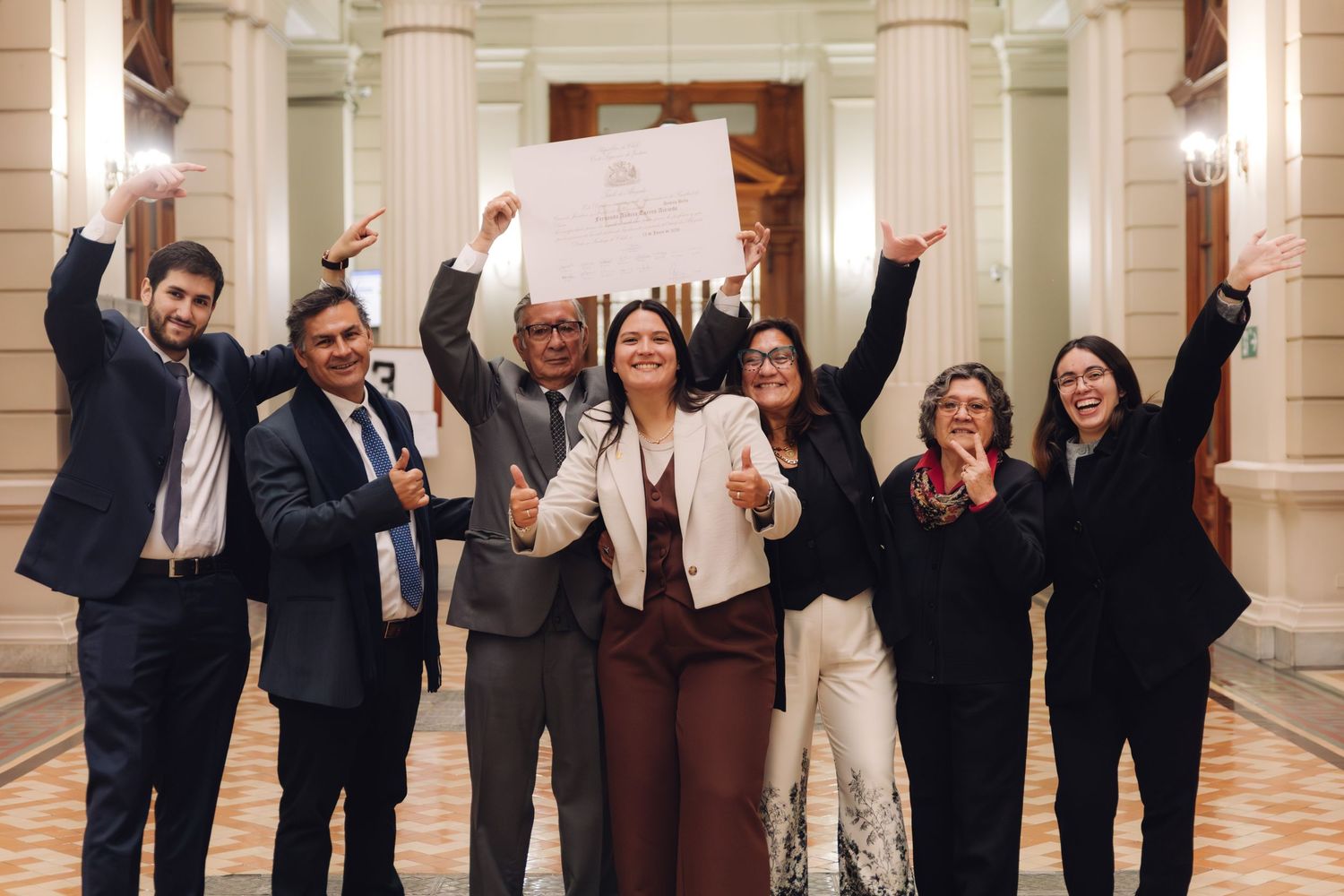 Joyful group of seven people celebrate with raised arms holding document in grand hall.