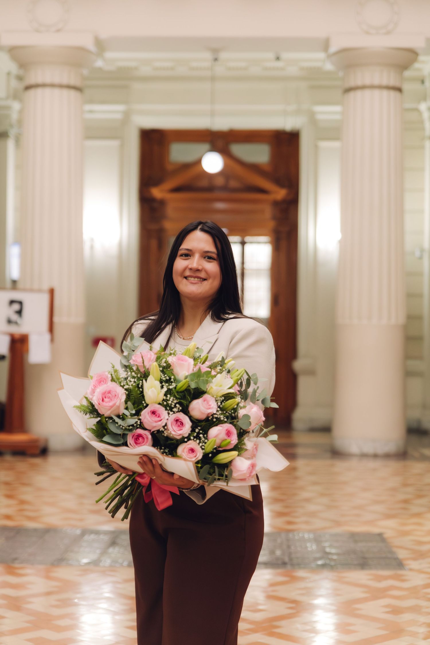 Smiling woman in white blazer holds large pink rose bouquet in classical columned hall.