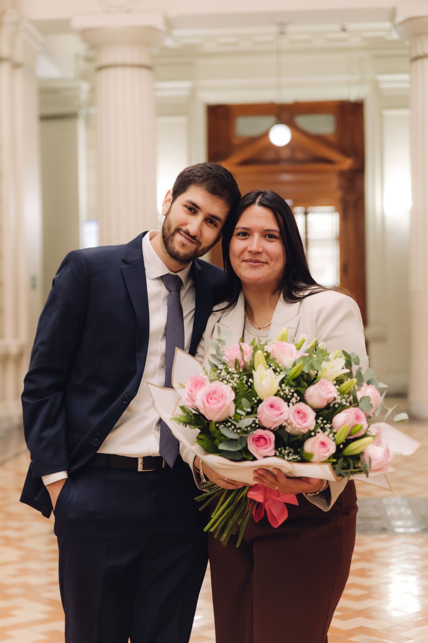 Man in navy suit and woman in white blazer pose with pink flower bouquet in formal hall.