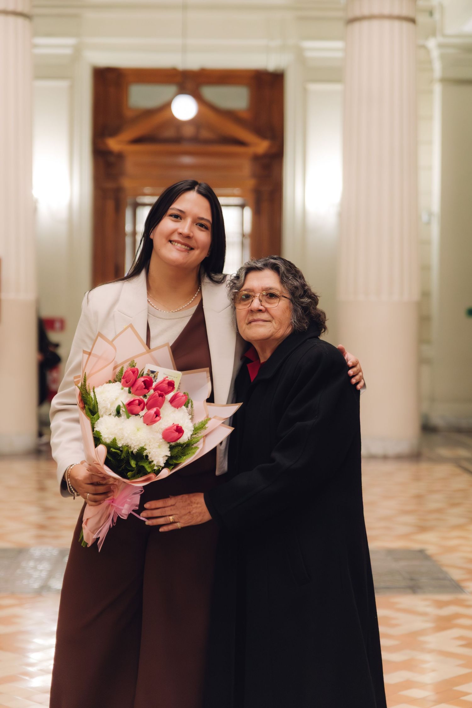 Tall woman in white blazer holds pink bouquet embracing elderly woman in black coat.