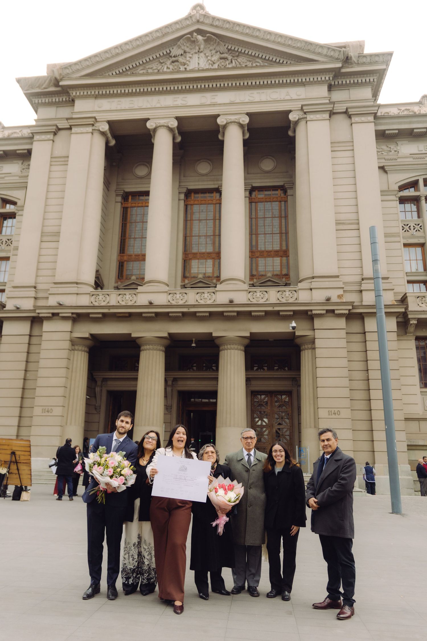 Group of seven people hold flowers and certificate outside neoclassical courthouse facade.