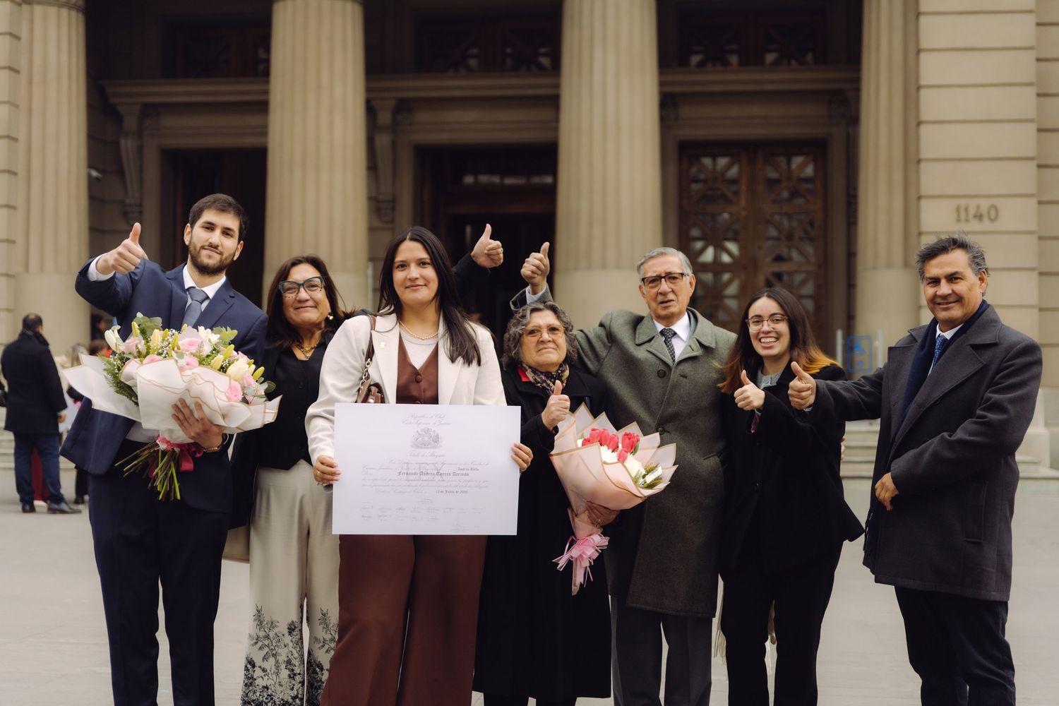 Group of seven people celebrating outdoors holding flowers and a diploma in front of a grand building.