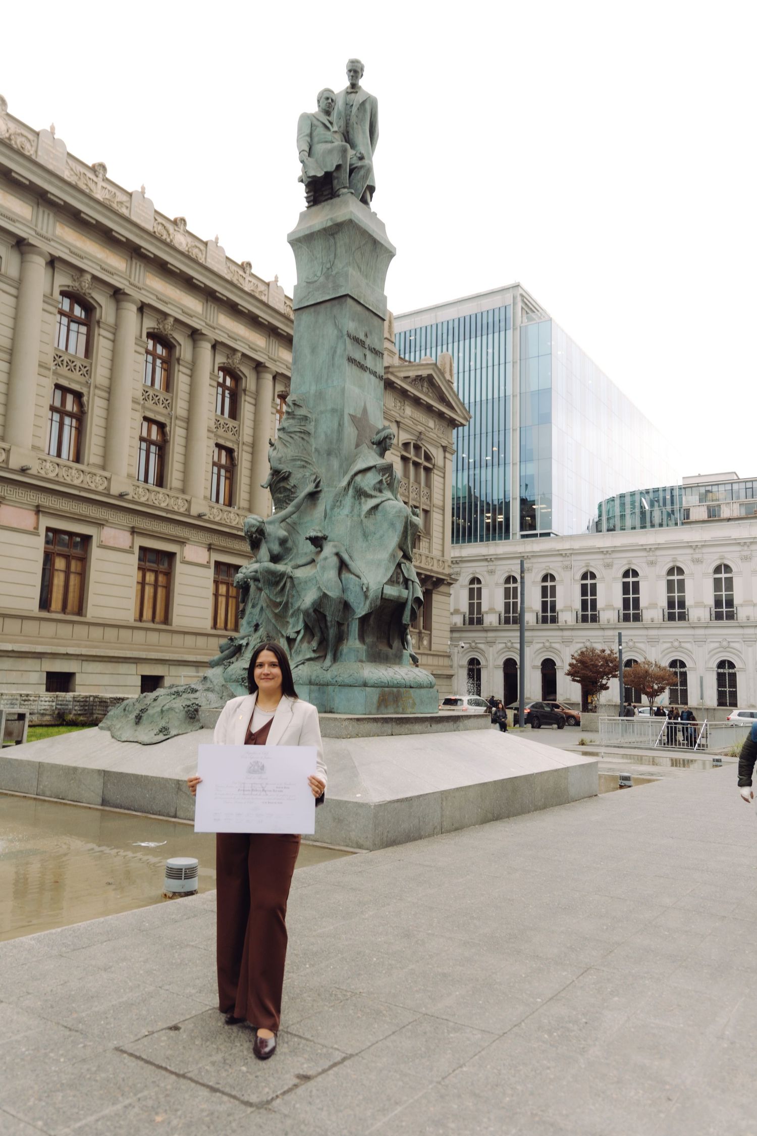 Young woman holding a white certificate sign standing in front of a bronze monument and neoclassical building.