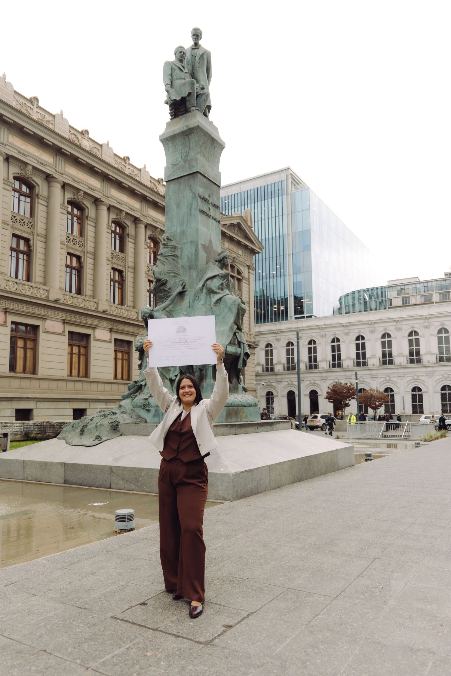Smiling woman triumphantly raises a diploma certificate in front of a historic monument and classical architecture.