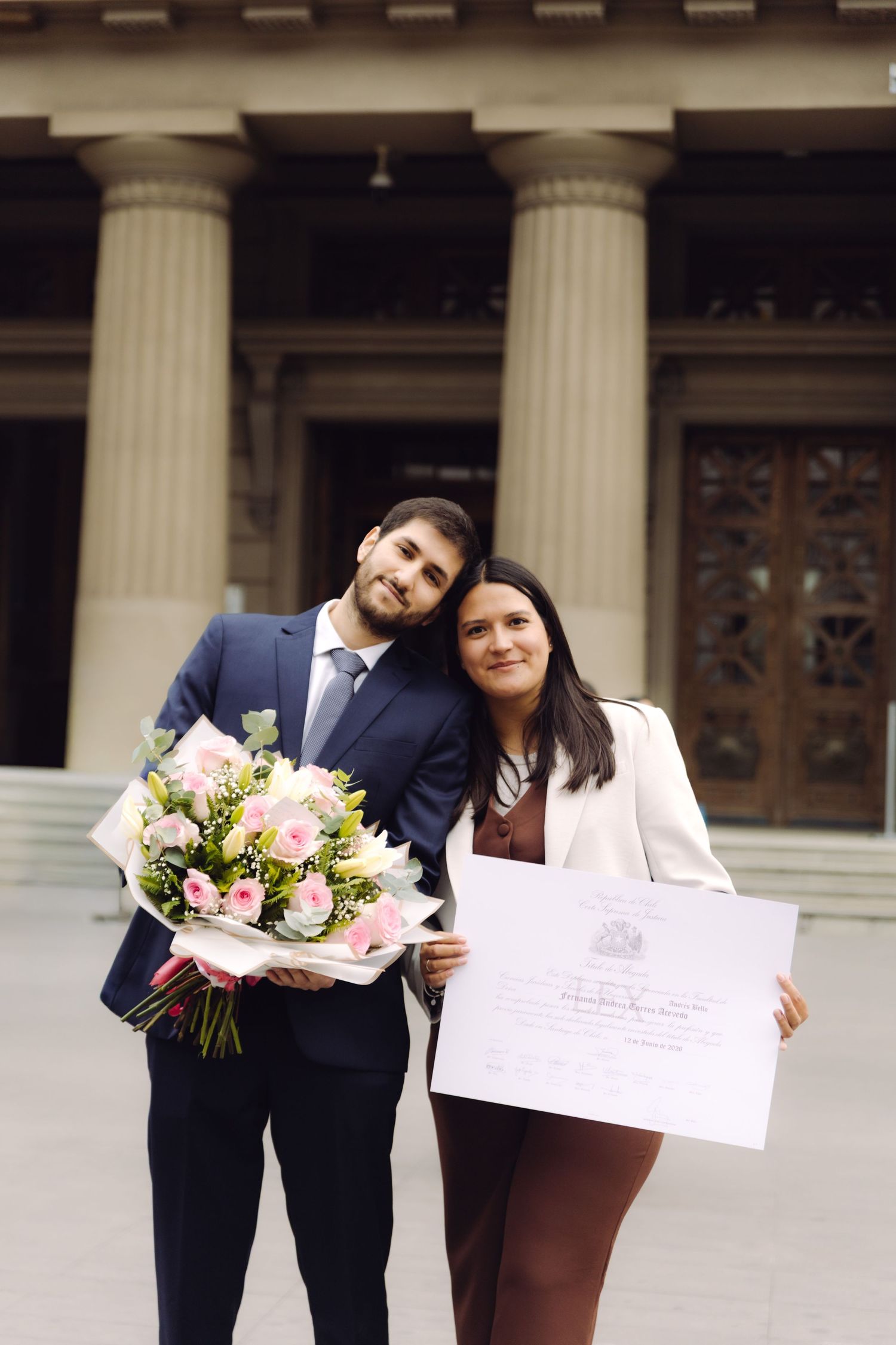 Man with bouquet and woman holding diploma pose proudly in front of a neoclassical building with columns.