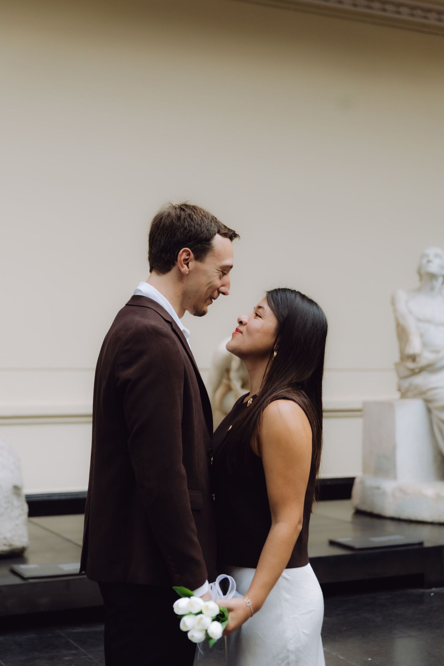 Smiling couple sharing a tender moment in a museum gallery with classical sculptures in the background.
